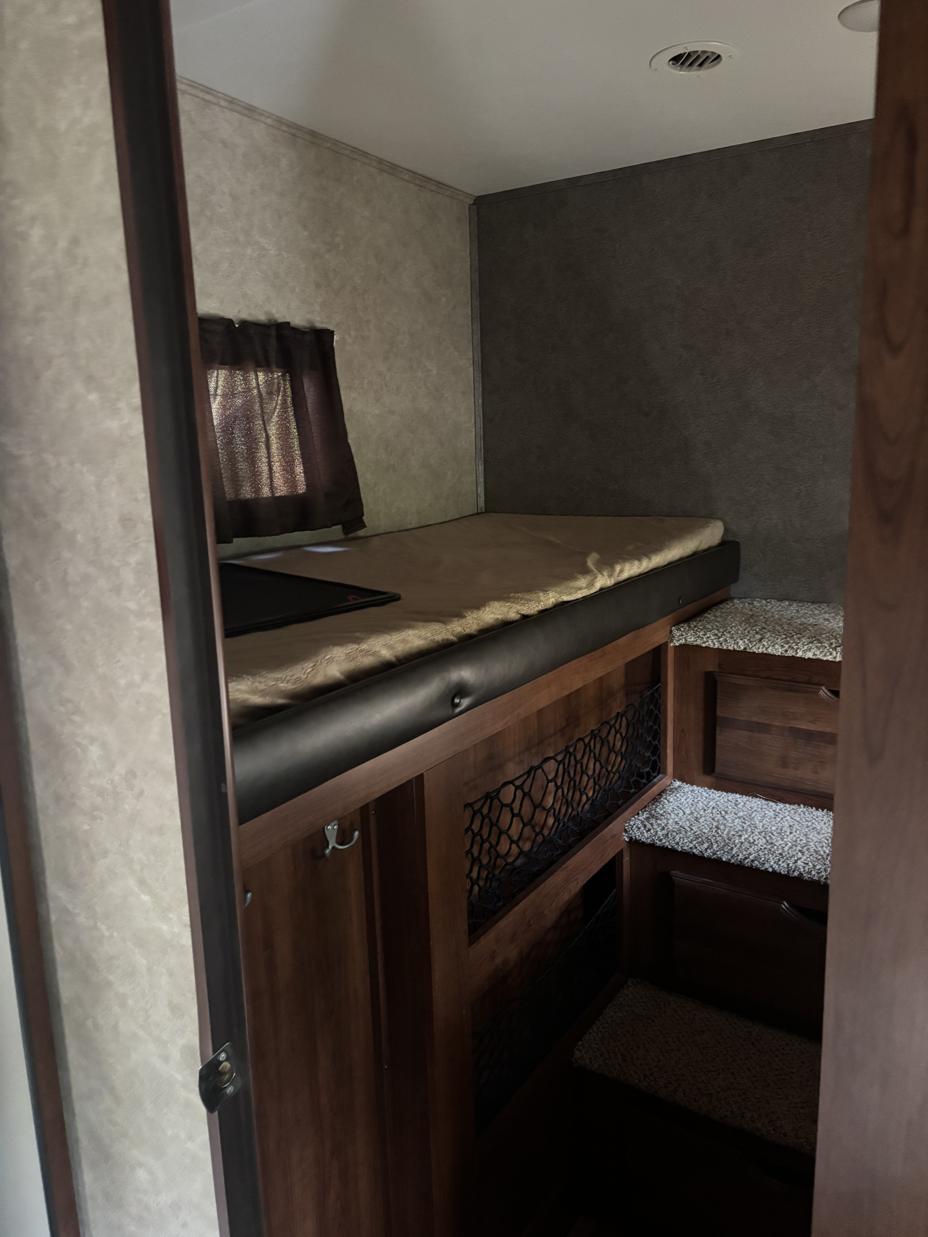 Inside an RV rental: a raised bed with light bedding, dark window curtains, built-in steps with carpet, and wood storage.. Jayco Eagle HT 2018