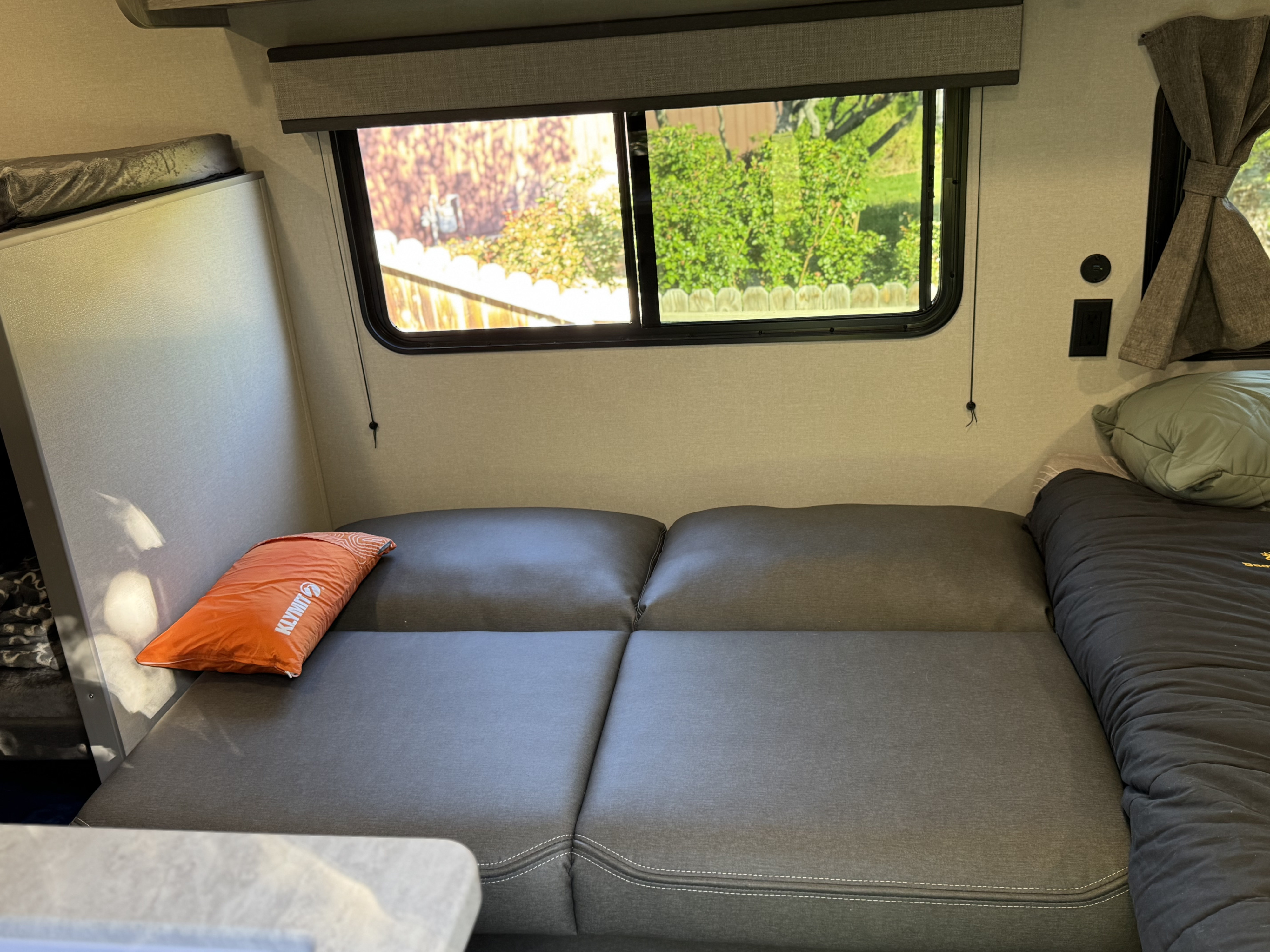 Cozy RV rental interior with grey cushions, Klymit pillow, rolled sleeping bag, and a view outside the window.. Forest river Aurora light 16BHX 2025