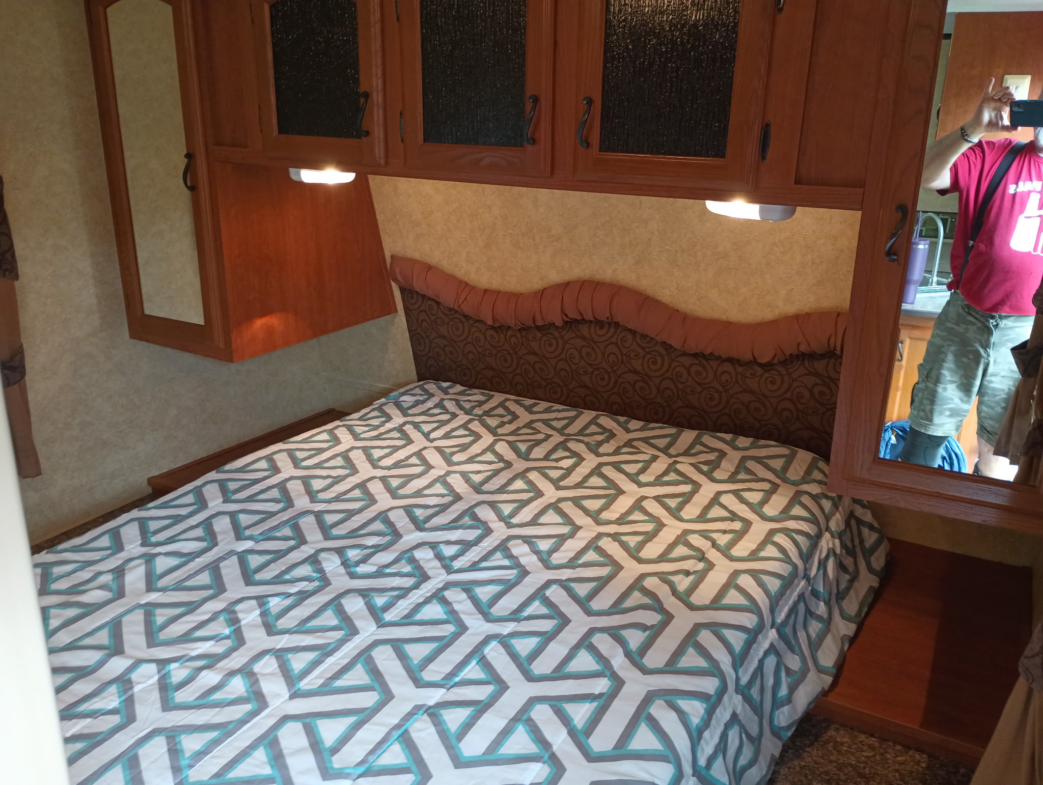 Inside an RV rental bedroom: comfy bed with patterned comforter, overhead storage, and mirror showing photographer.. Forest River Tracer Ultra Lite 2012