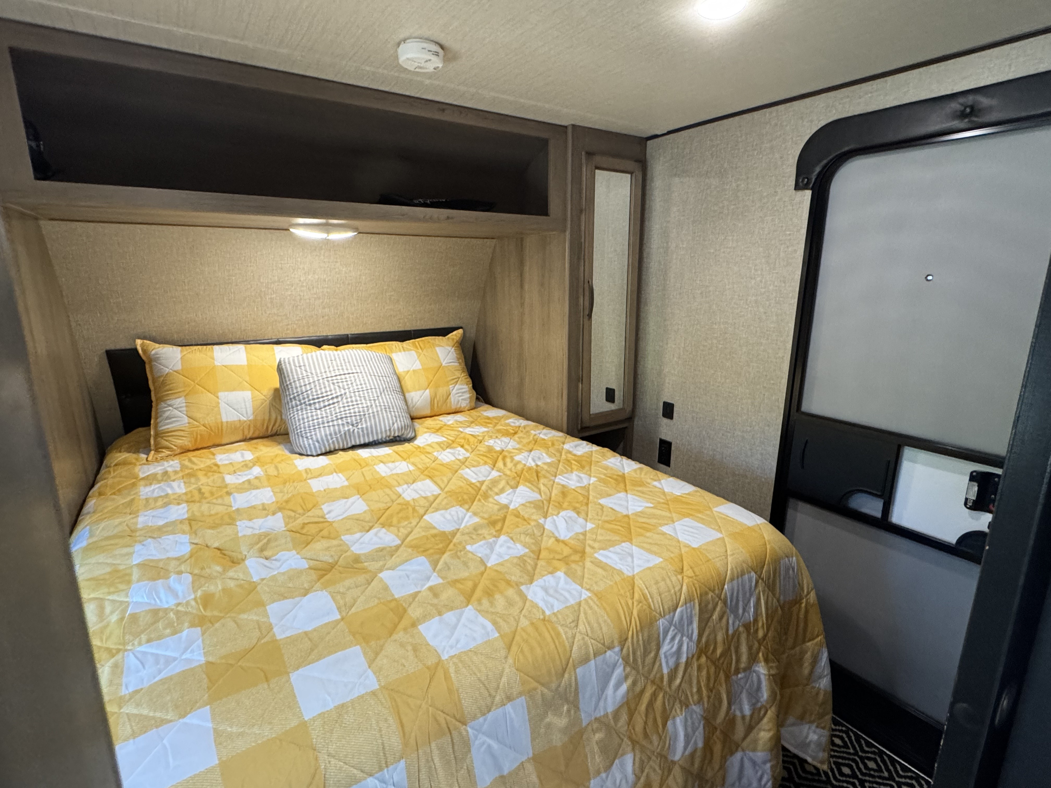Cozy RV rental bedroom features a comfy bed with a bright yellow checkered quilt, overhead storage, and a convenient door.. Grand Design Transcend Xplor 2020