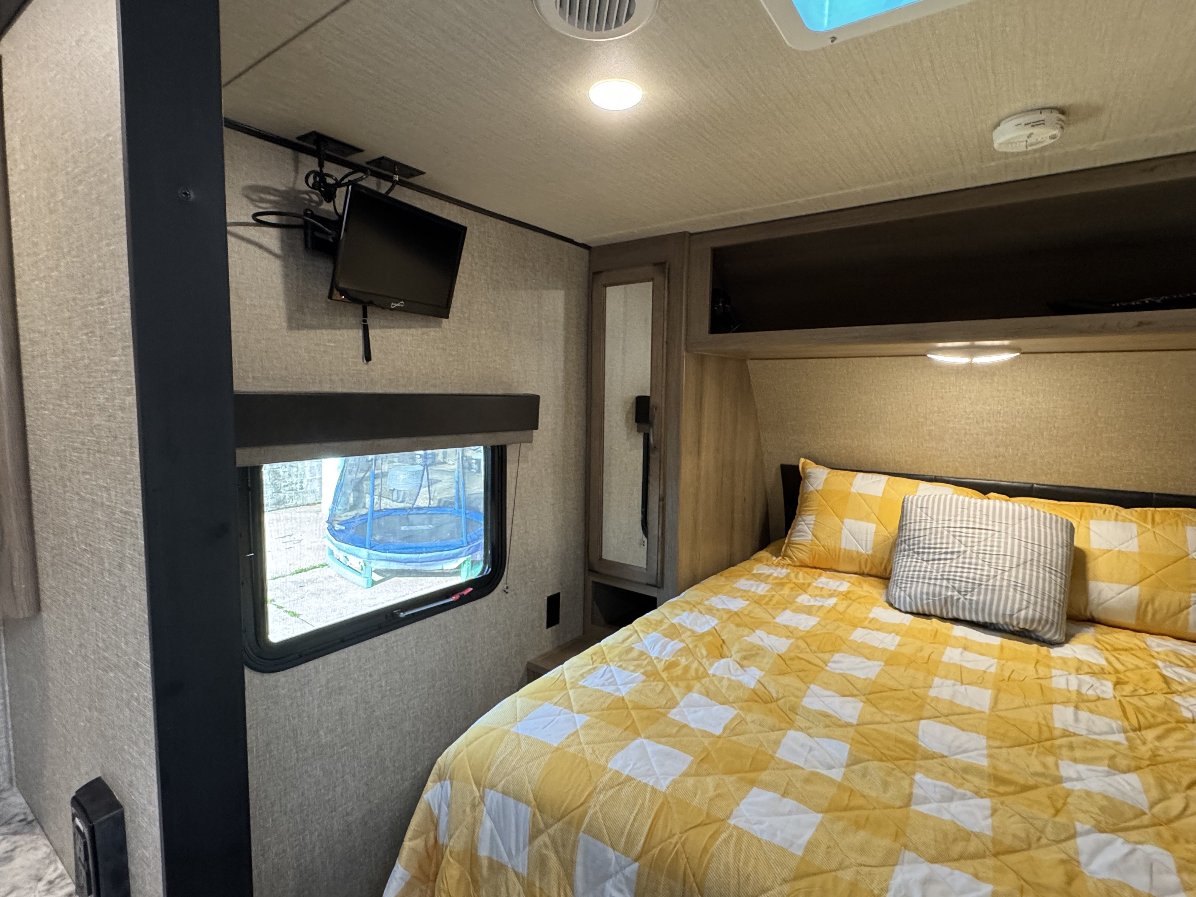 Cozy RV rental bedroom with checkered bed, window view of a trampoline, and wall-mounted TV.. Grand Design Transcend Xplor 2020