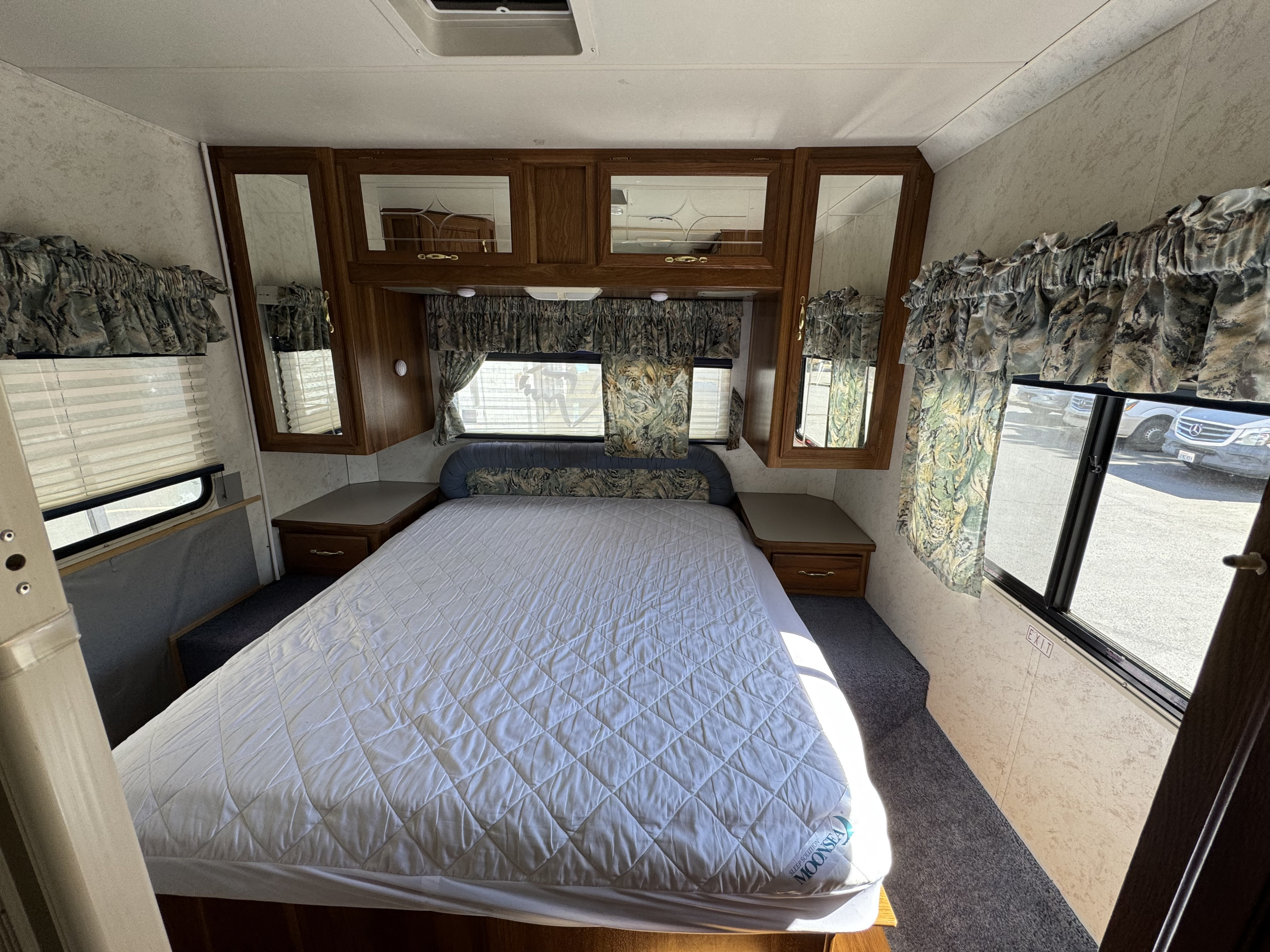 Explore this cozy RV bedroom, ideal for your RV rental. Features a large bed, wood cabinets, and scenic windows.. Thor Motor Coach Four Winds 1999