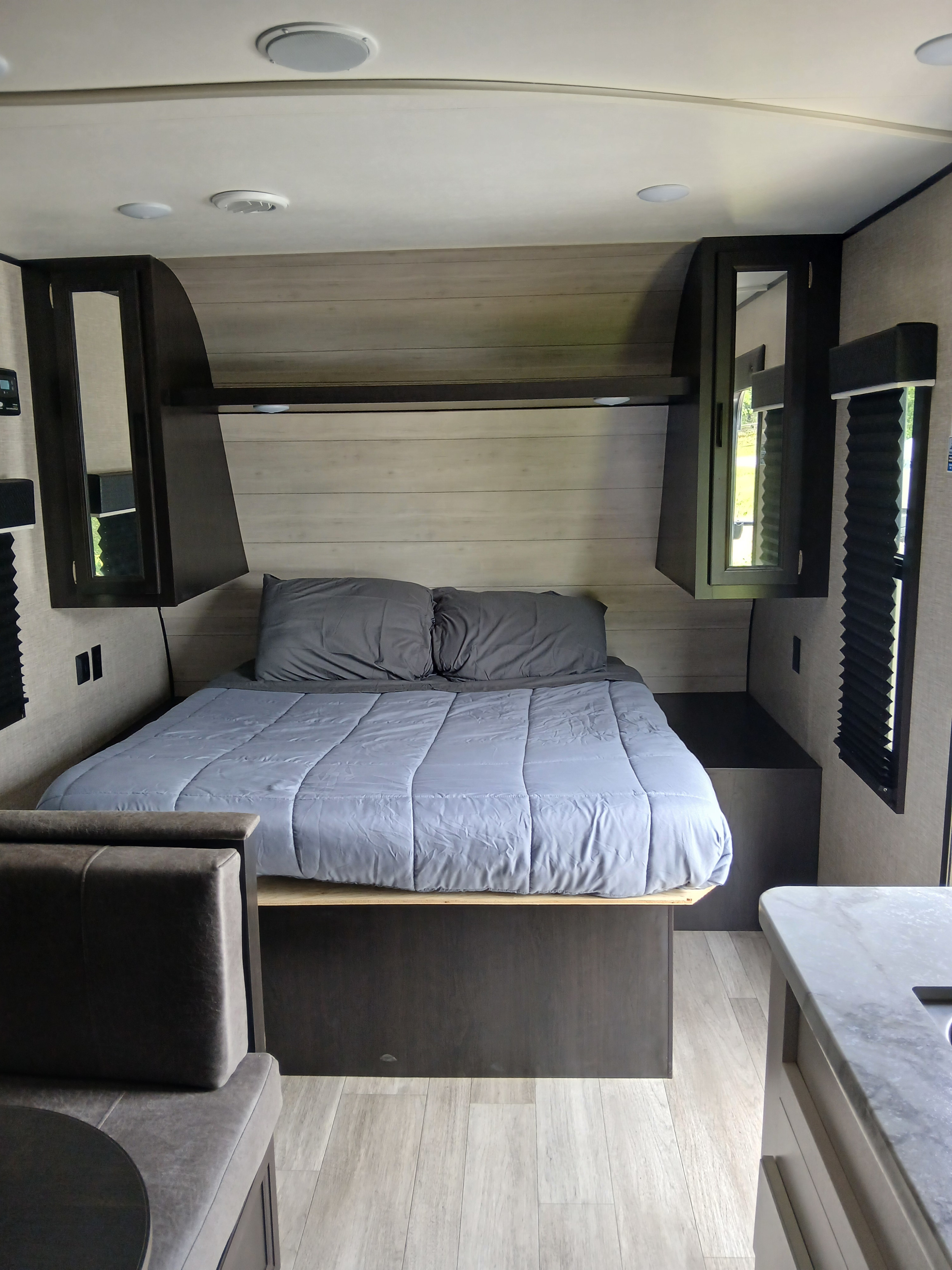 Interior of an RV rental bedroom showing a gray bed, overhead cabinets, and dinette seating on wood-look flooring.. Jayco 2022
