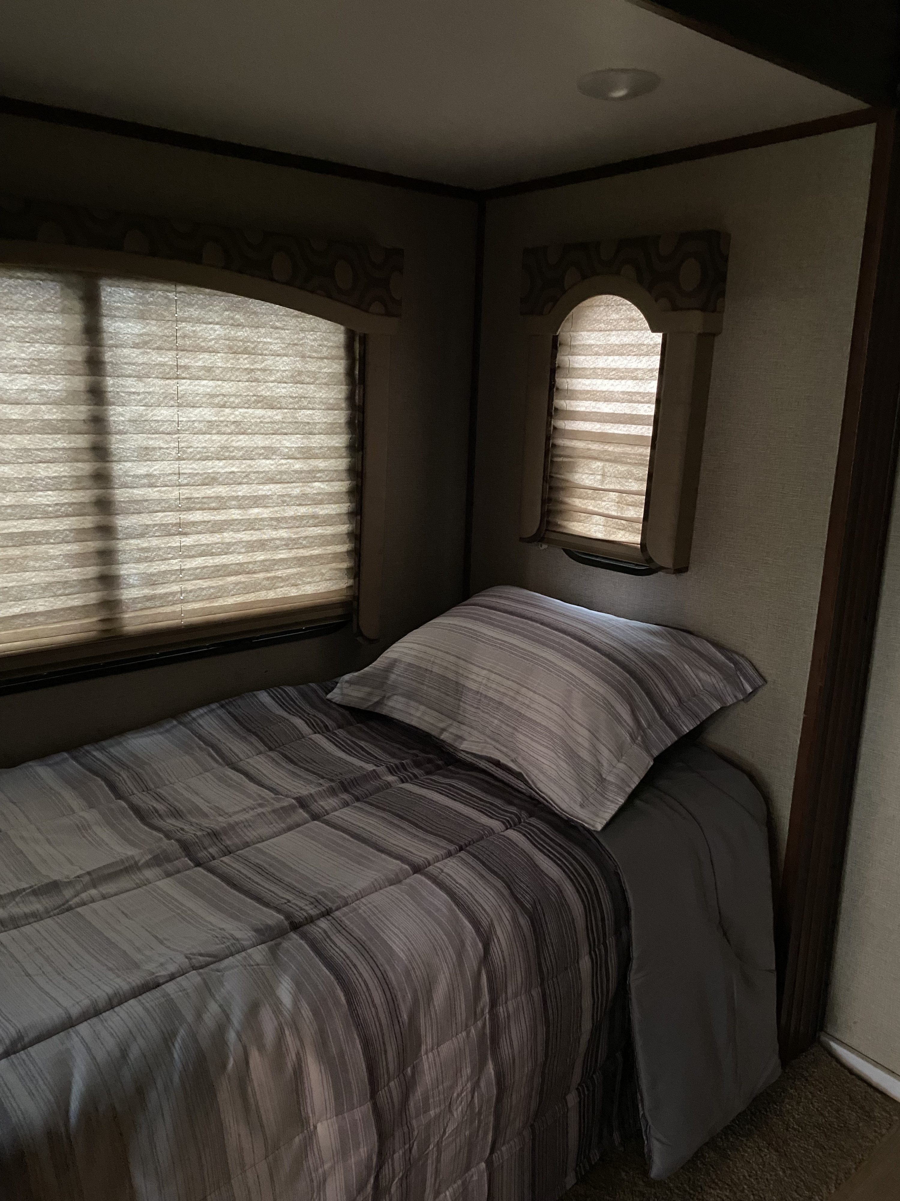 An RV rental bedroom features a striped bed, pillow, and two windows with drawn pleated blinds.. Keystone RV Avalanche 2017