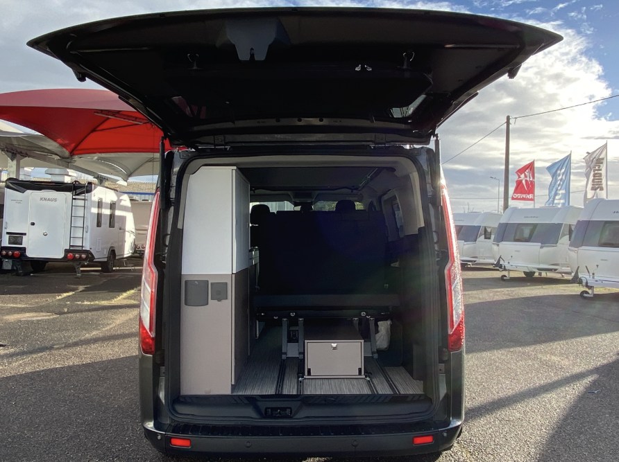 Looking into an RV rental camper van from the rear, showing its cabinet, bed, and storage. Other RVs are visible outside.. BURSTNER COPA 500 2023