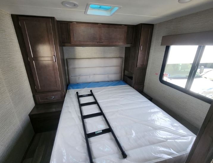 This RV rental bedroom features a new bed with a plastic-wrapped mattress, wooden cabinets, and a window. A ladder rests on the bed.. Winnebago Spirit 2023