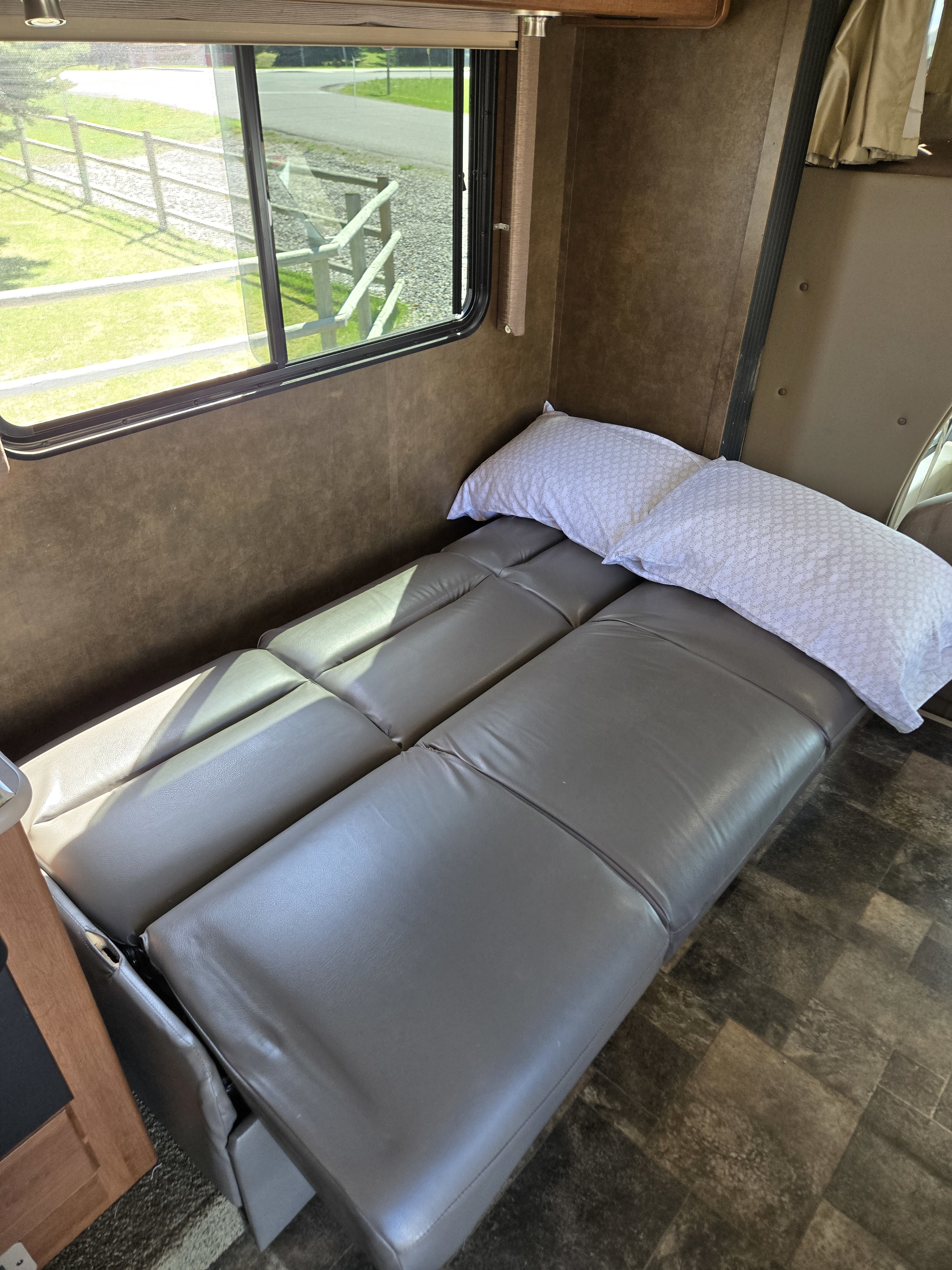 An RV rental interior with a grey sofa/bed, pillows, and a bright window view of a fenced road.. Tioga Ranger 2014
