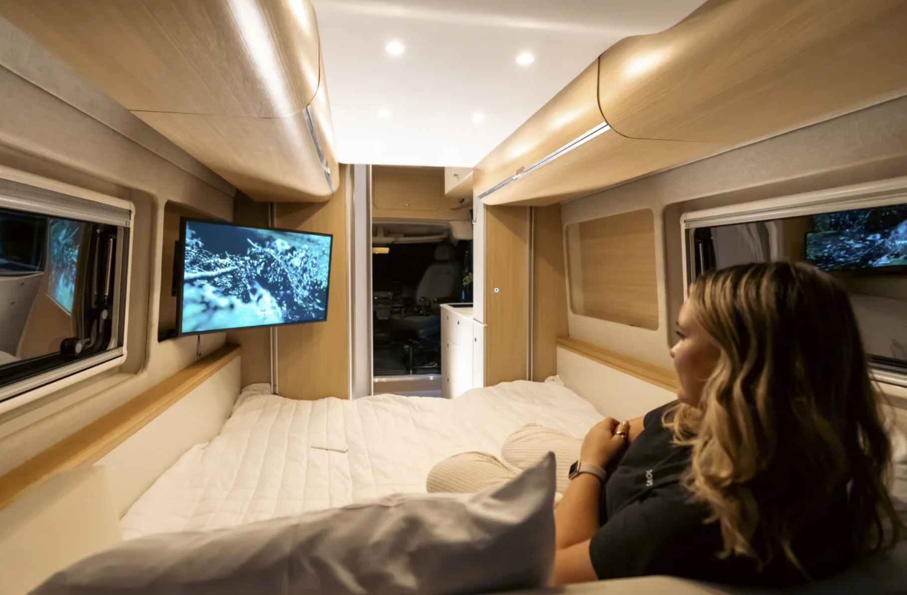 Inside a modern RV rental, a woman with long hair lies on the comfy bed watching TV. Wooden walls & ceiling add warmth.. Dodge Ram Promaster 2025