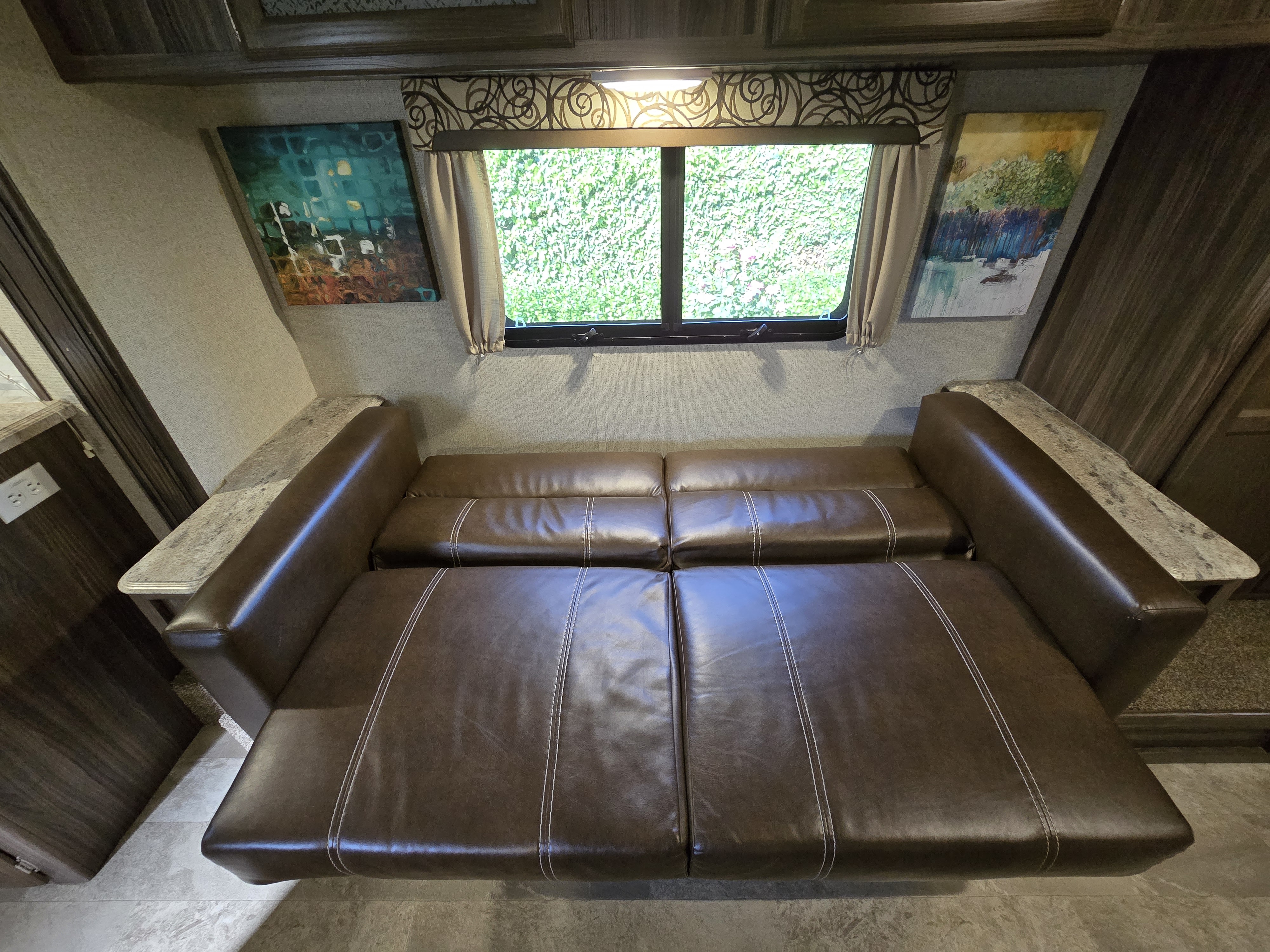 This RV rental features a versatile brown sofa that converts into a bed, flanked by abstract art and a window with green views.. Coachmen Apex 2017
