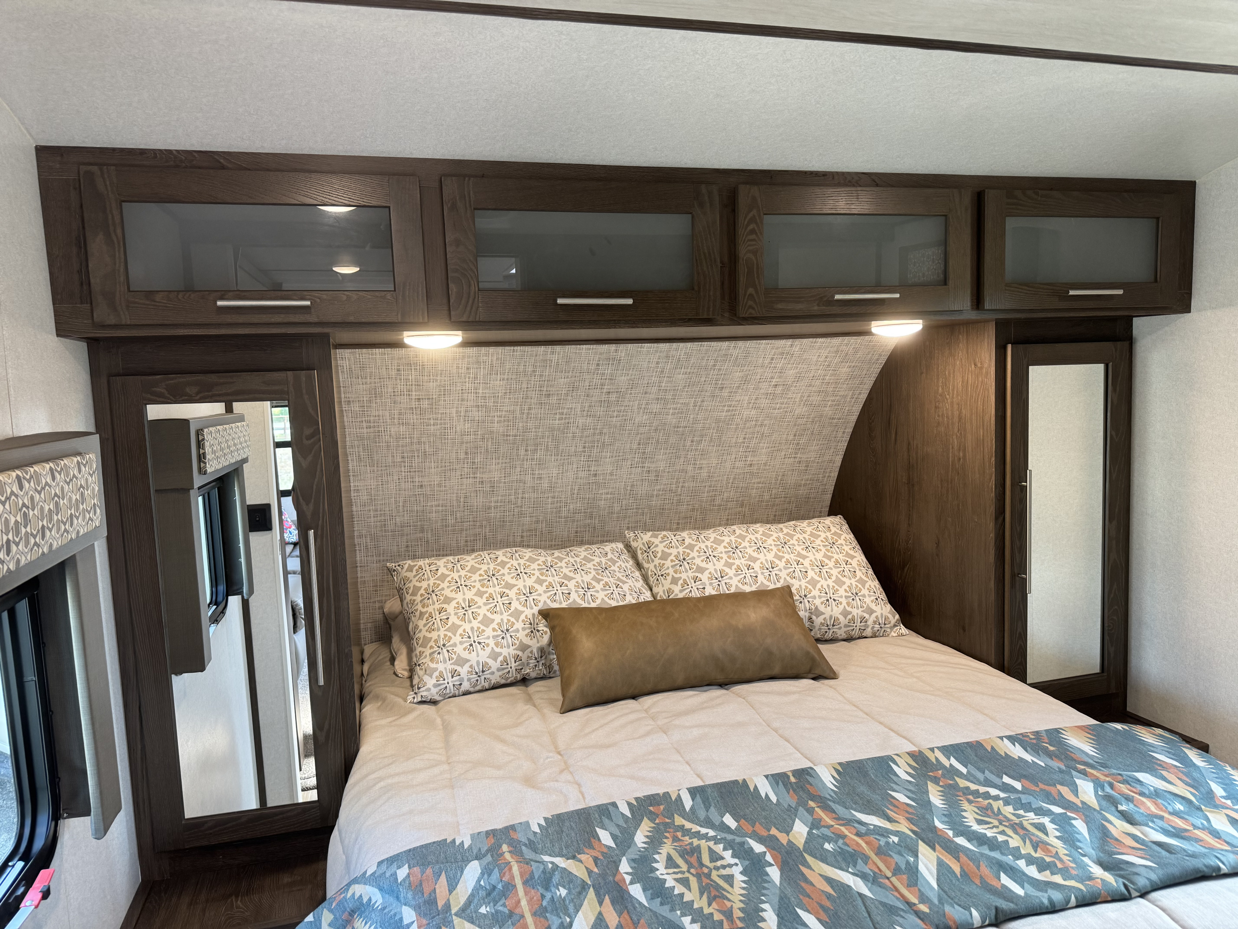 RV rental bedroom showcasing a bed, dark wood overhead cabinets, upholstered headboard, and mirrored wardrobes.. Wildwood Heritage Glen 22RBHL 2019