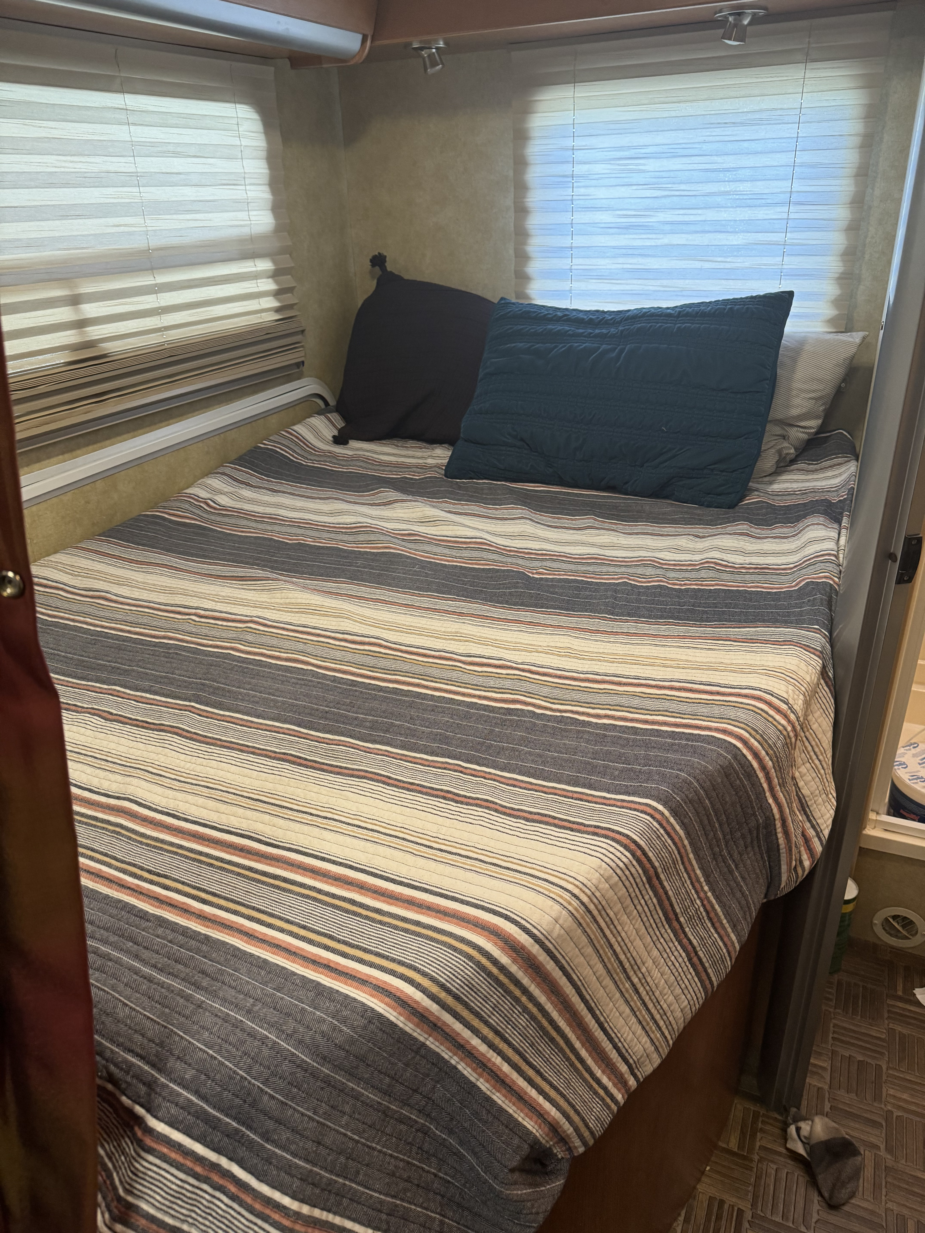 This cozy RV rental bedroom features a bed with a striped comforter and pillows, flanked by windows with blinds.. Itasca Navion 2007