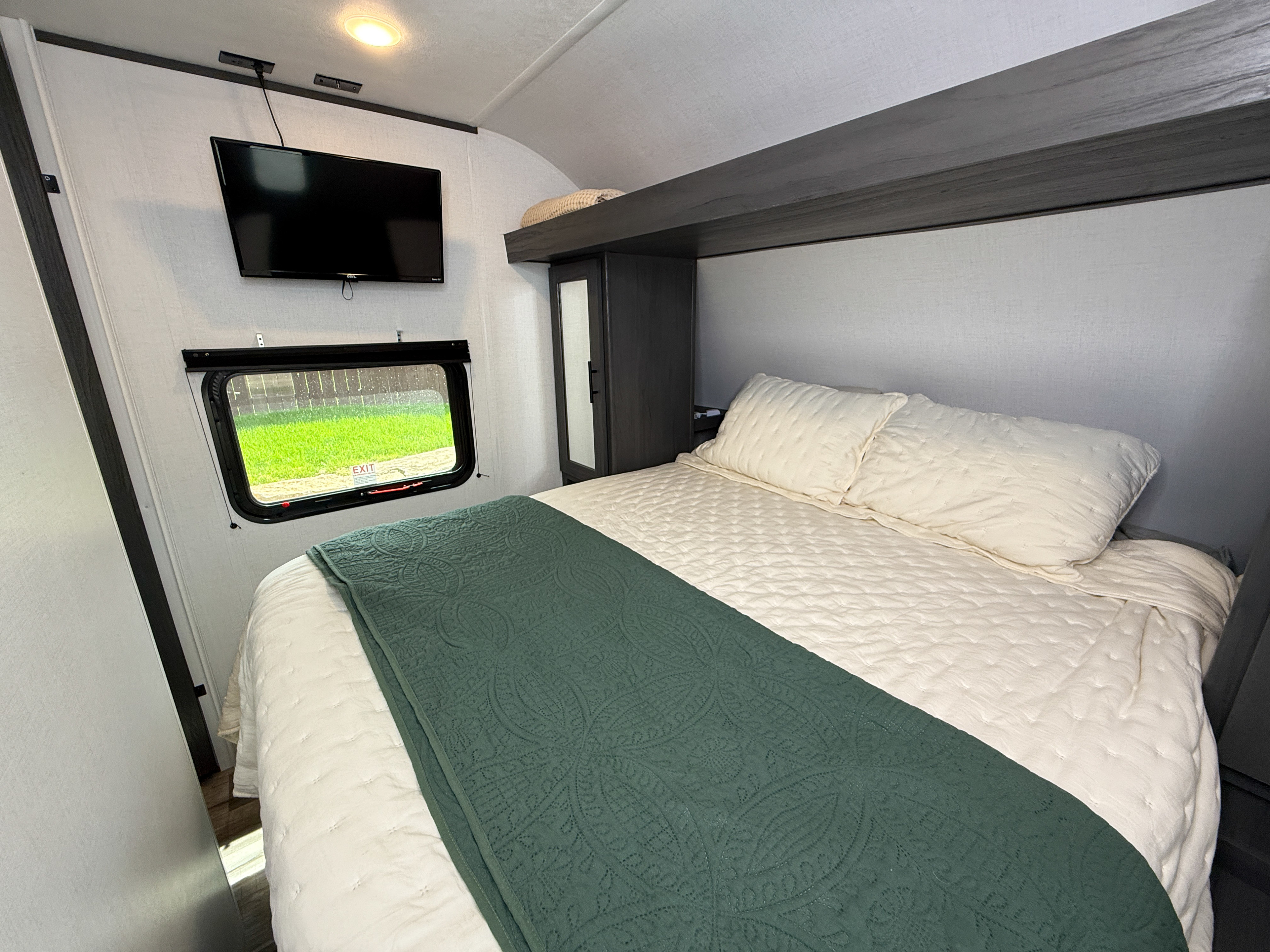 Spacious RV rental bedroom featuring a comfortable bed with a green blanket, wall-mounted TV, and window view of grass.. Keystone Dutchmen Colorado 29BHC 2025
