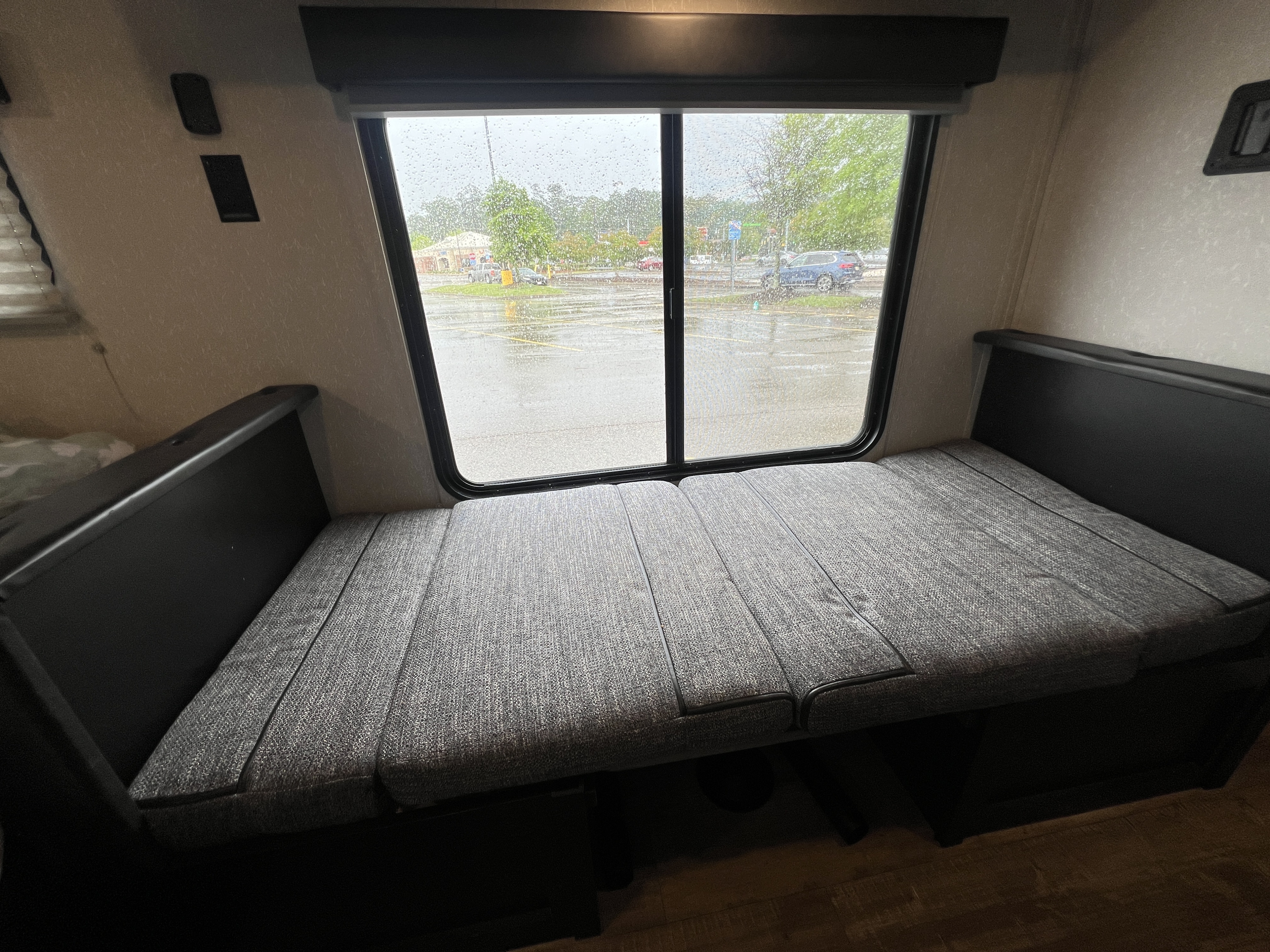 RV rental dinette/bed setup with grey cushions & a window view of a rainy parking lot. Cozy interior.. Forest River Cherokee Wolf Pup 2021