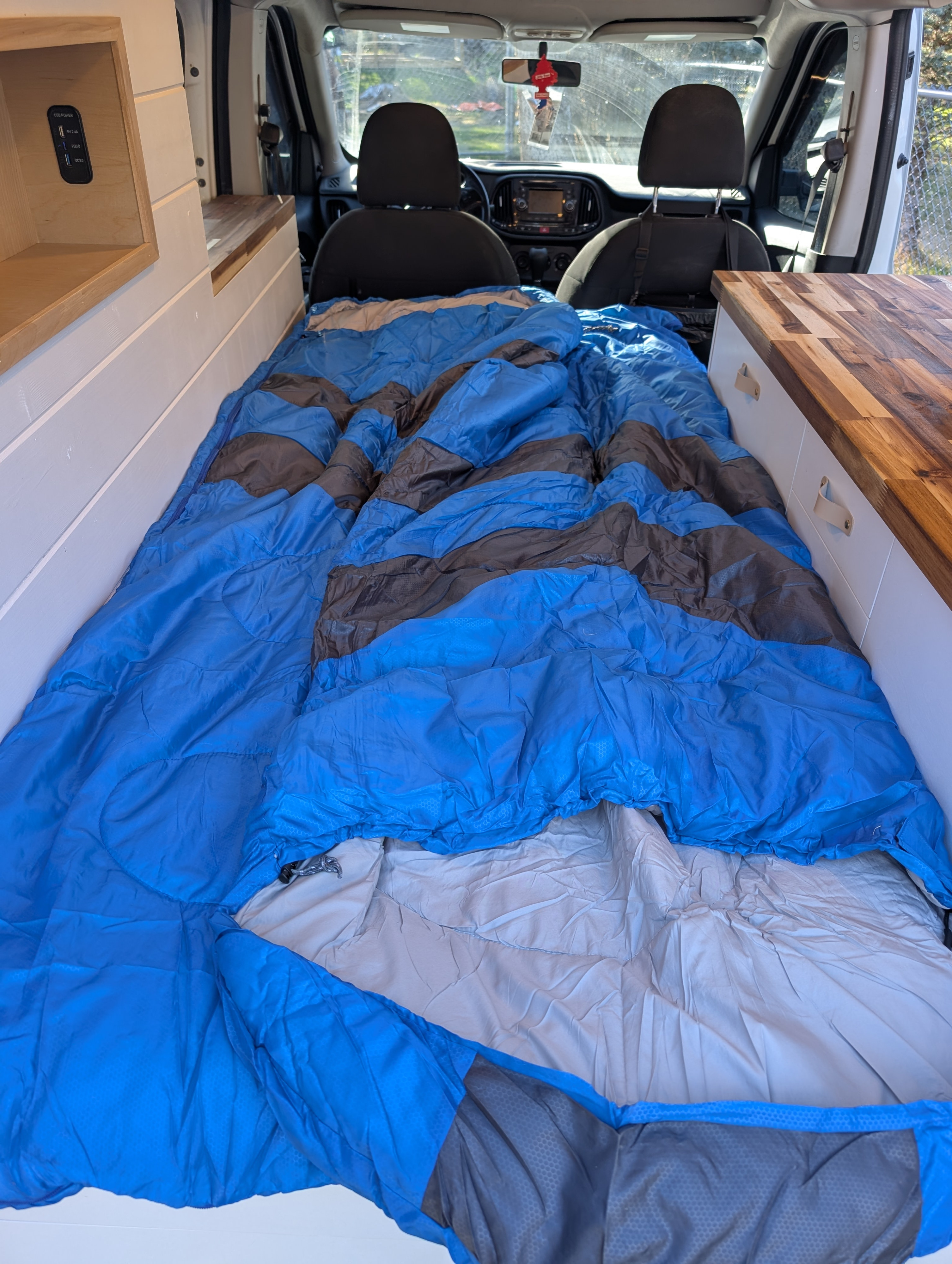Cozy RV rental interior: a comfortable bed with blue sleeping bag, custom wood shelving & counters.. Dodge Sprinter Van 2015