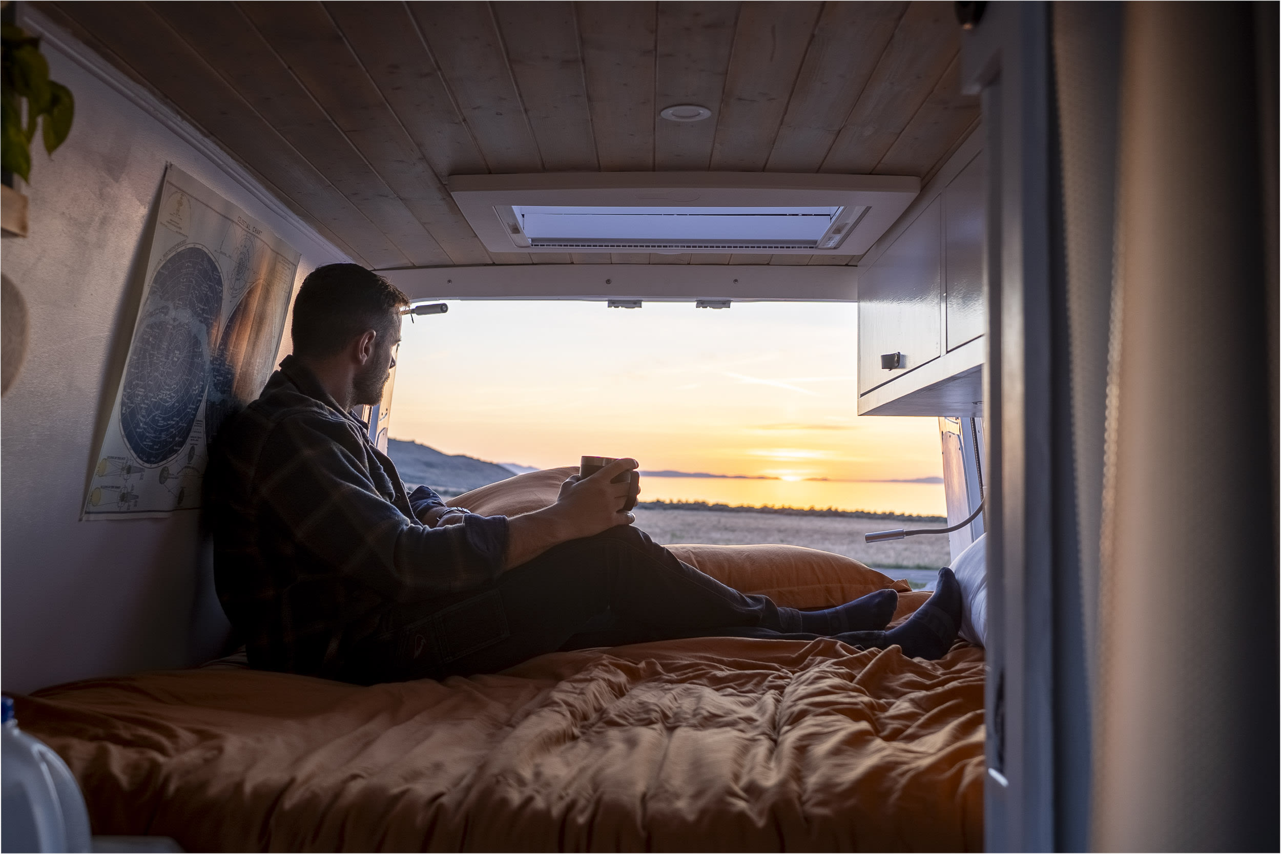 Enjoy stunning sunset views from a cozy RV rental! Man relaxes on the bed of his camper van, looking out at a lake and hills.. Freightliner Sprinter 2017