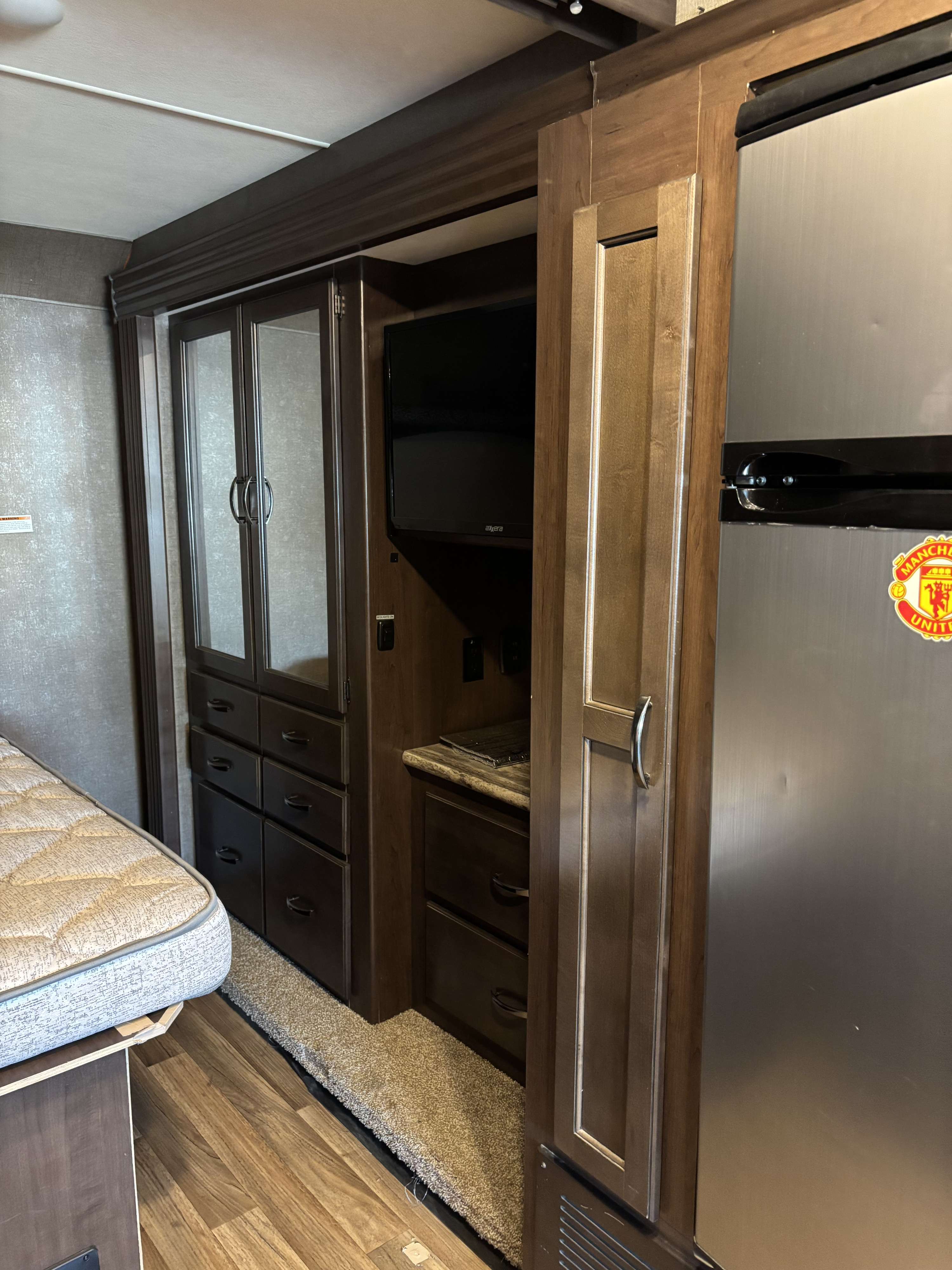 Compact RV rental bedroom with dark wood storage, mounted TV, and a stainless steel refrigerator.. Thor A.C.E. 2018