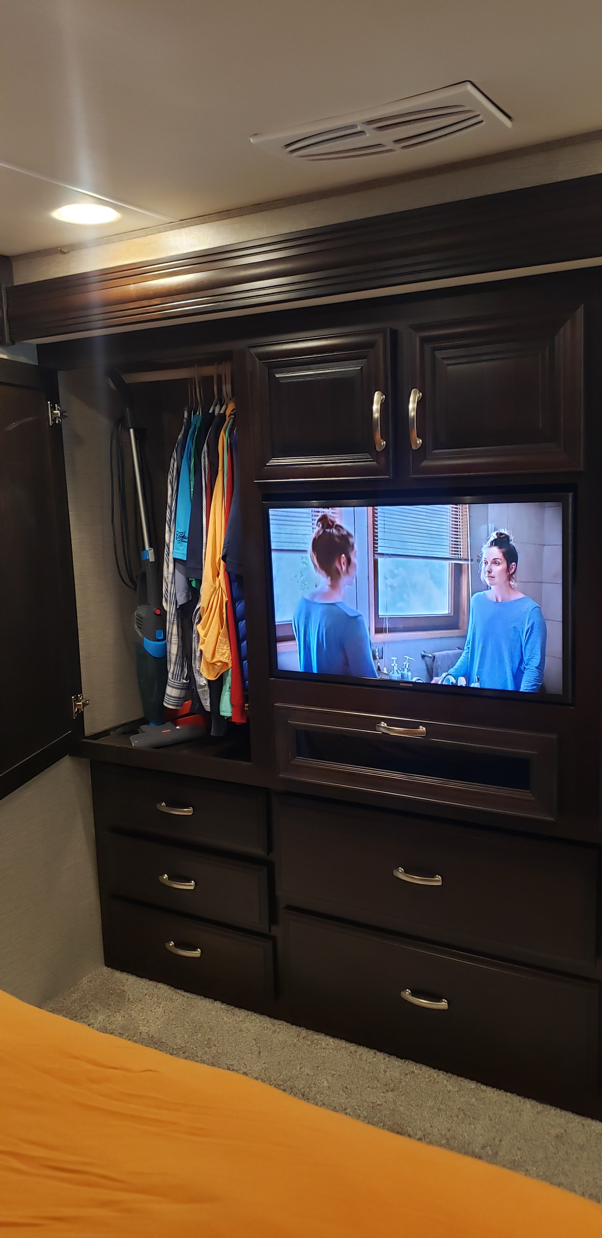 This RV rental bedroom features a built-in TV, wardrobe with hanging clothes, and dresser drawers, offering smart storage and comfort.. Berkshire Berkshire Motorhome 2019