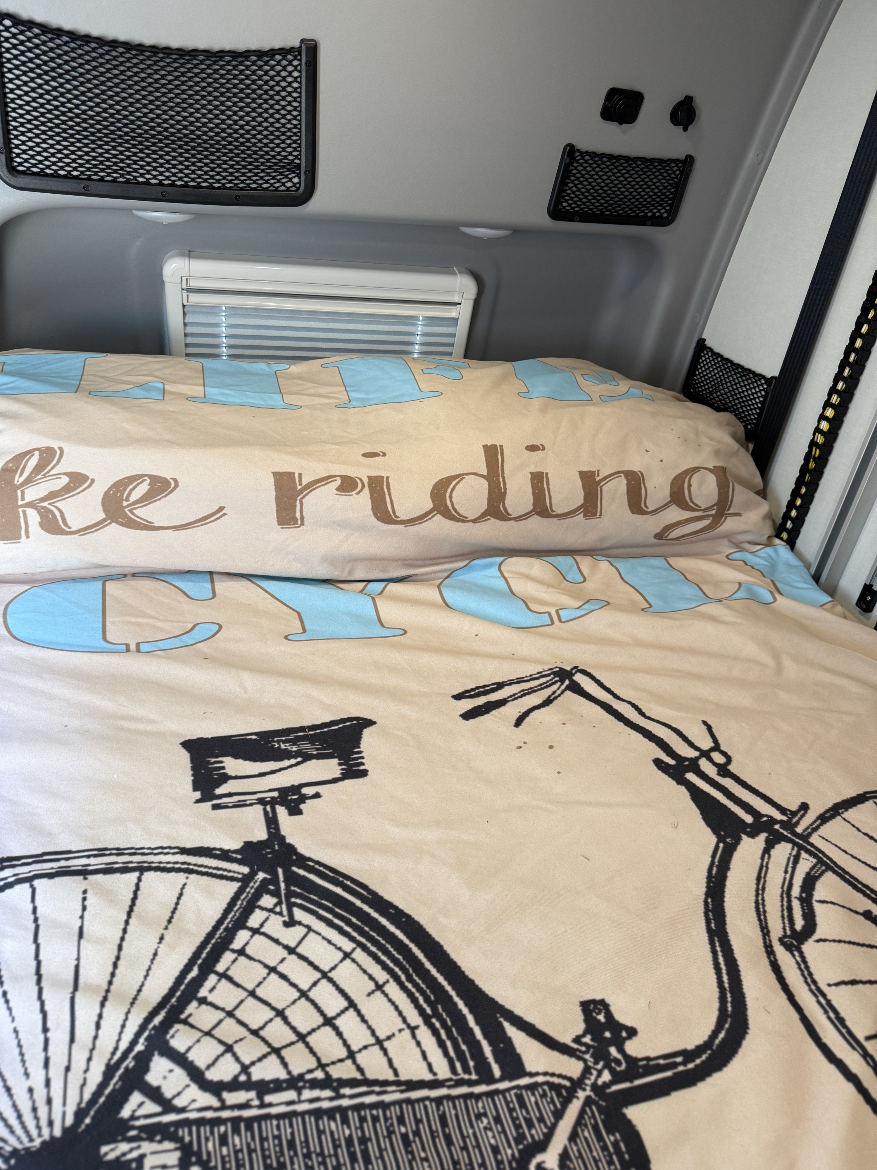 RV rental interior featuring a cozy bed with a bicycle-themed duvet, storage nets, & window blinds.. Winnebago Revel 2022