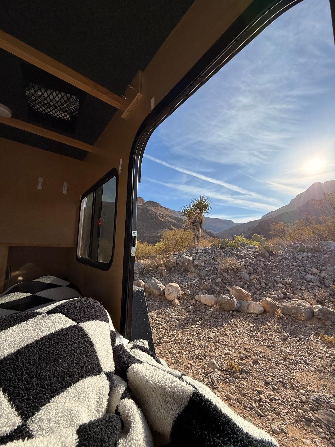 Cozy RV rental with a checkered blanket open to a stunning desert mountain landscape. Sun shines on a Yucca in the rocky terrain.. Outbound Outbound Extreme 2019
