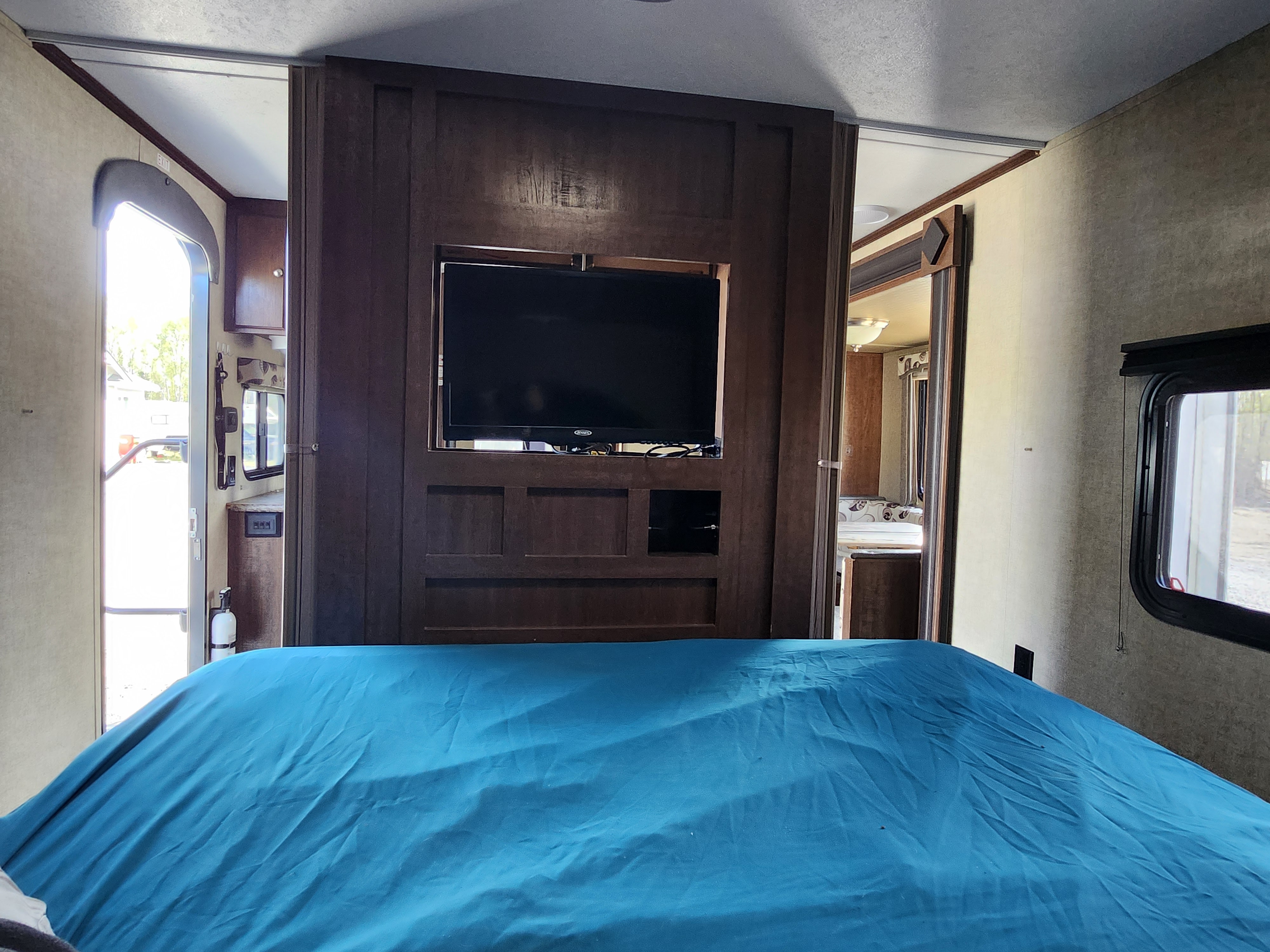 Interior of an RV rental: a bedroom with a blue bed, wall-mounted TV, and open doors leading to the exterior and living area.. Keystone RV Hideout 2015