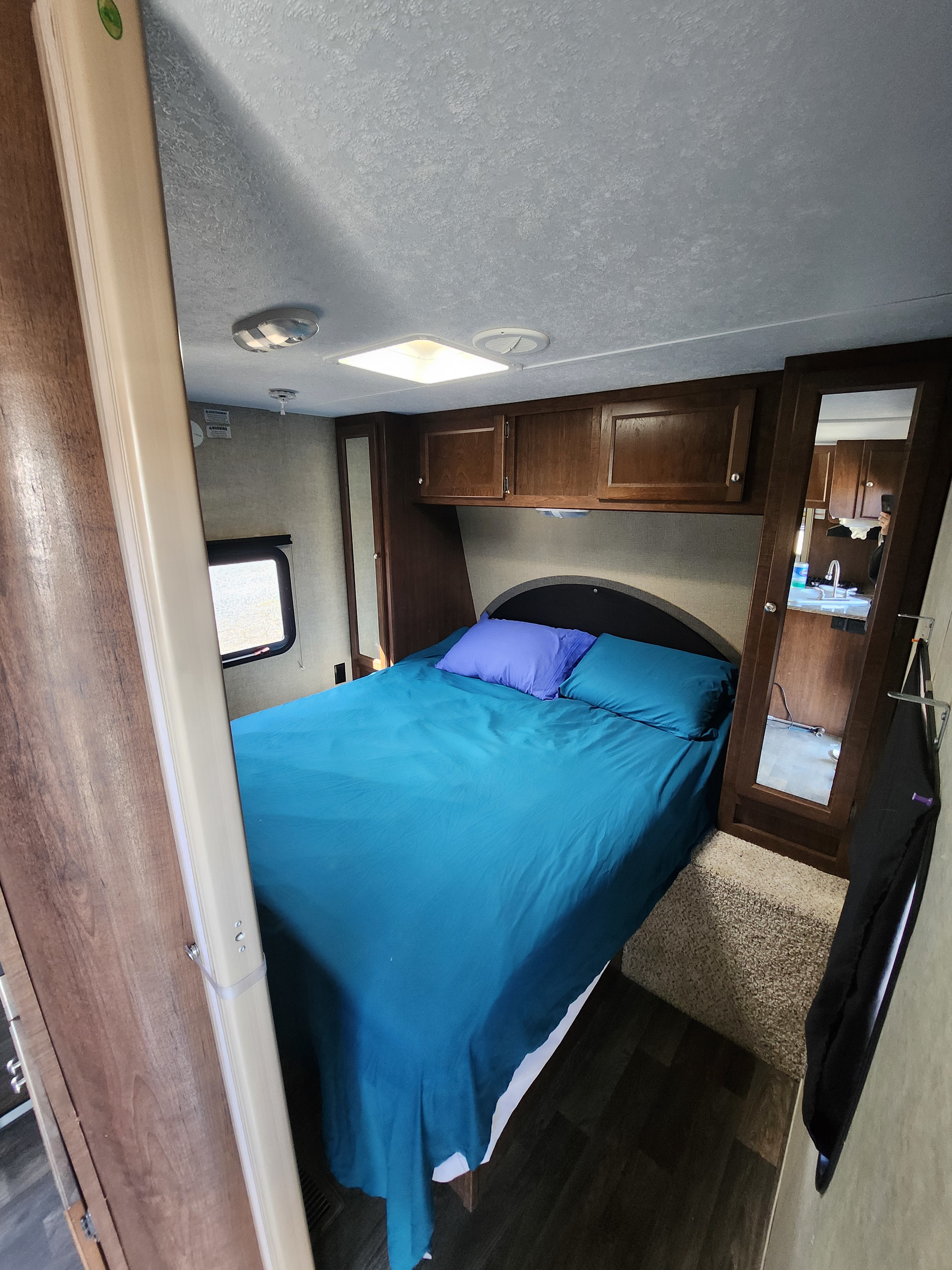 Cozy RV rental bedroom featuring a bed with blue bedding, overhead cabinets, and a mirror reflecting the kitchen. Ideal for travel.. Keystone RV Hideout 2015
