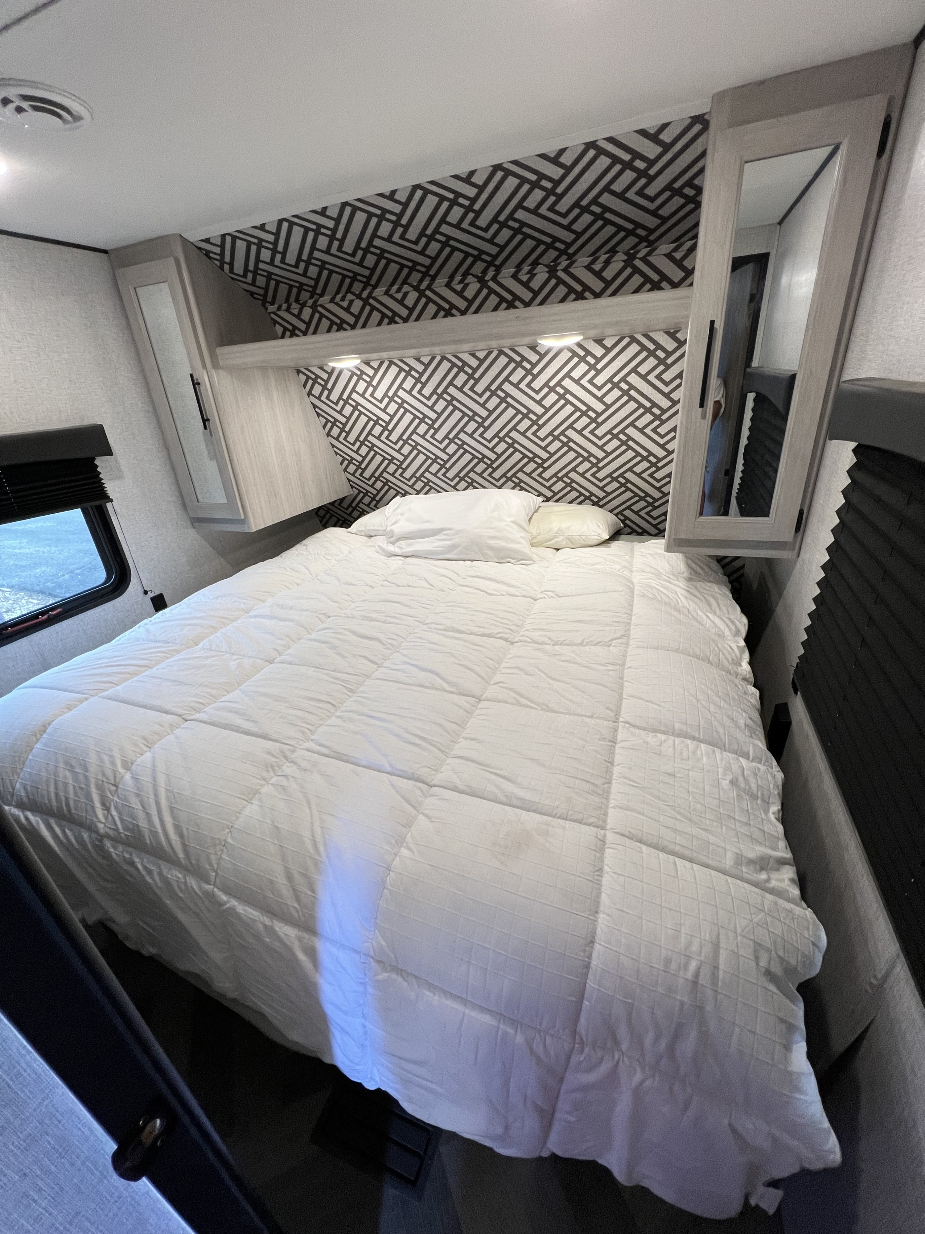 An inviting RV rental bedroom featuring a comfy white bed, overhead storage with mirrors, geometric wallpaper, and reading lights.. Crossroads RV Zinger 2024