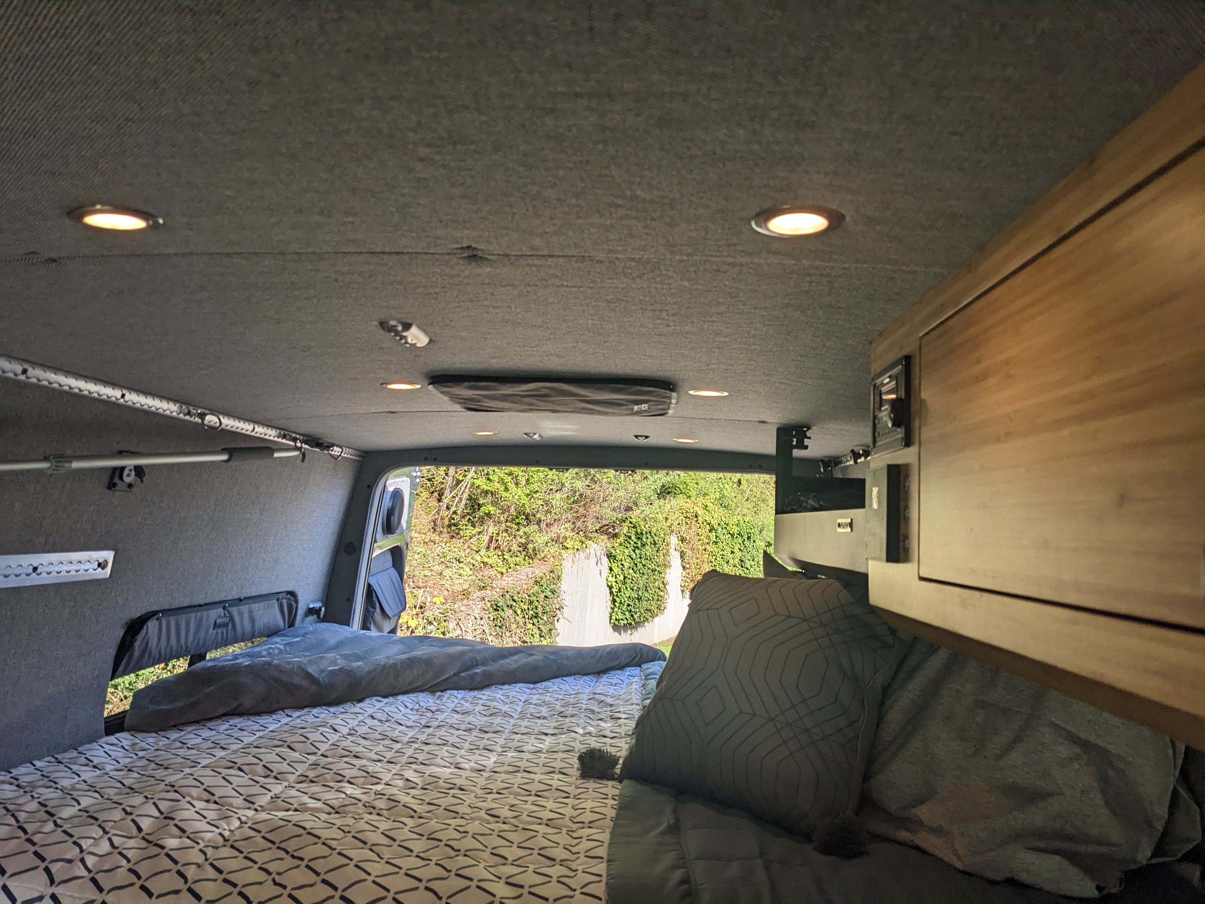 Inside a modern RV rental, a comfy bed with patterned sheets fills the view. Grey walls, overhead lights, & wood cabinets.. Mercedes Sprinter 2022