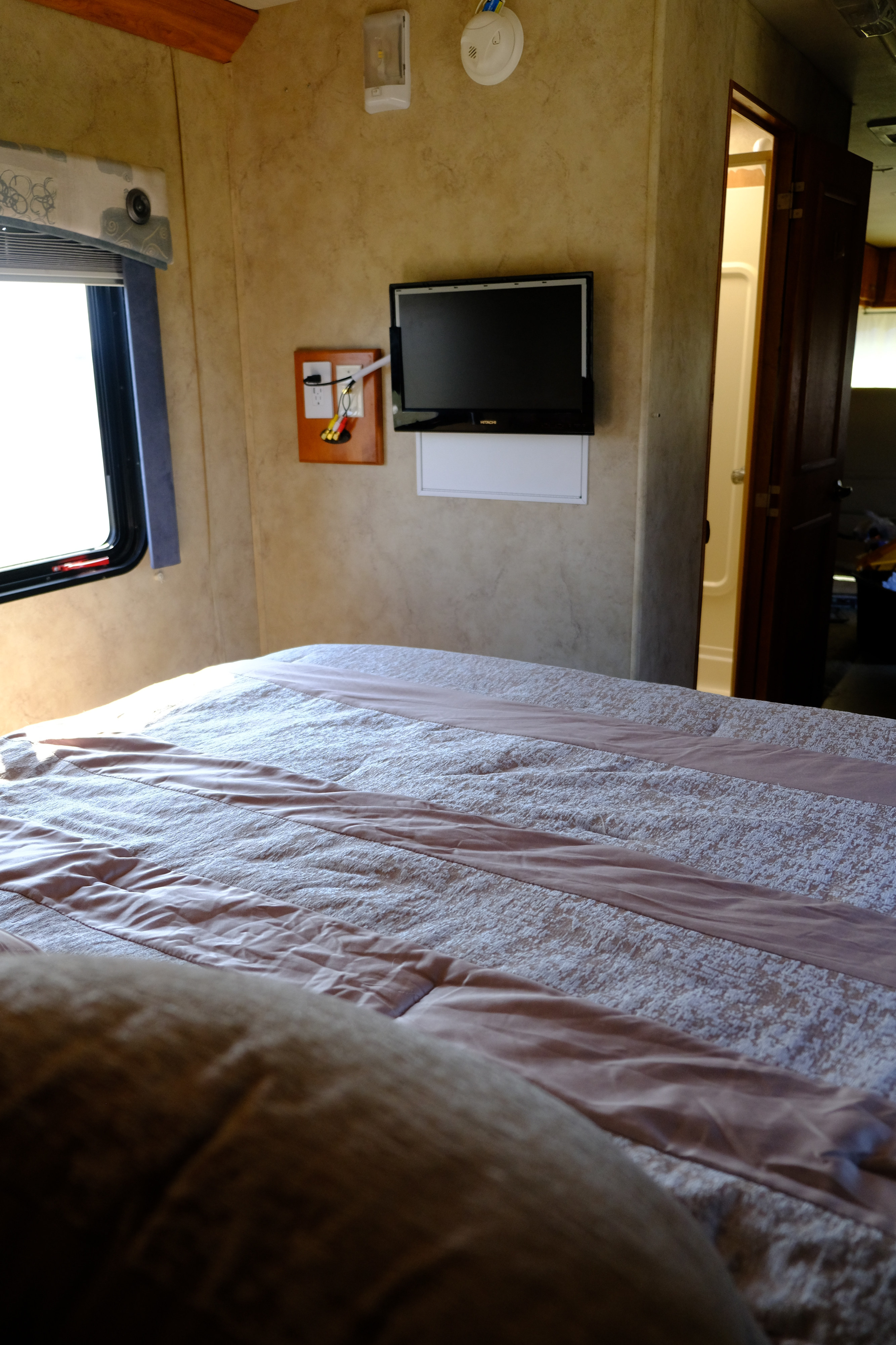 Inside an RV rental, showing a bedroom with a made bed, wall-mounted TV, window with blinds, and a partial view of the bathroom.. Gulf Stream Independence 2006