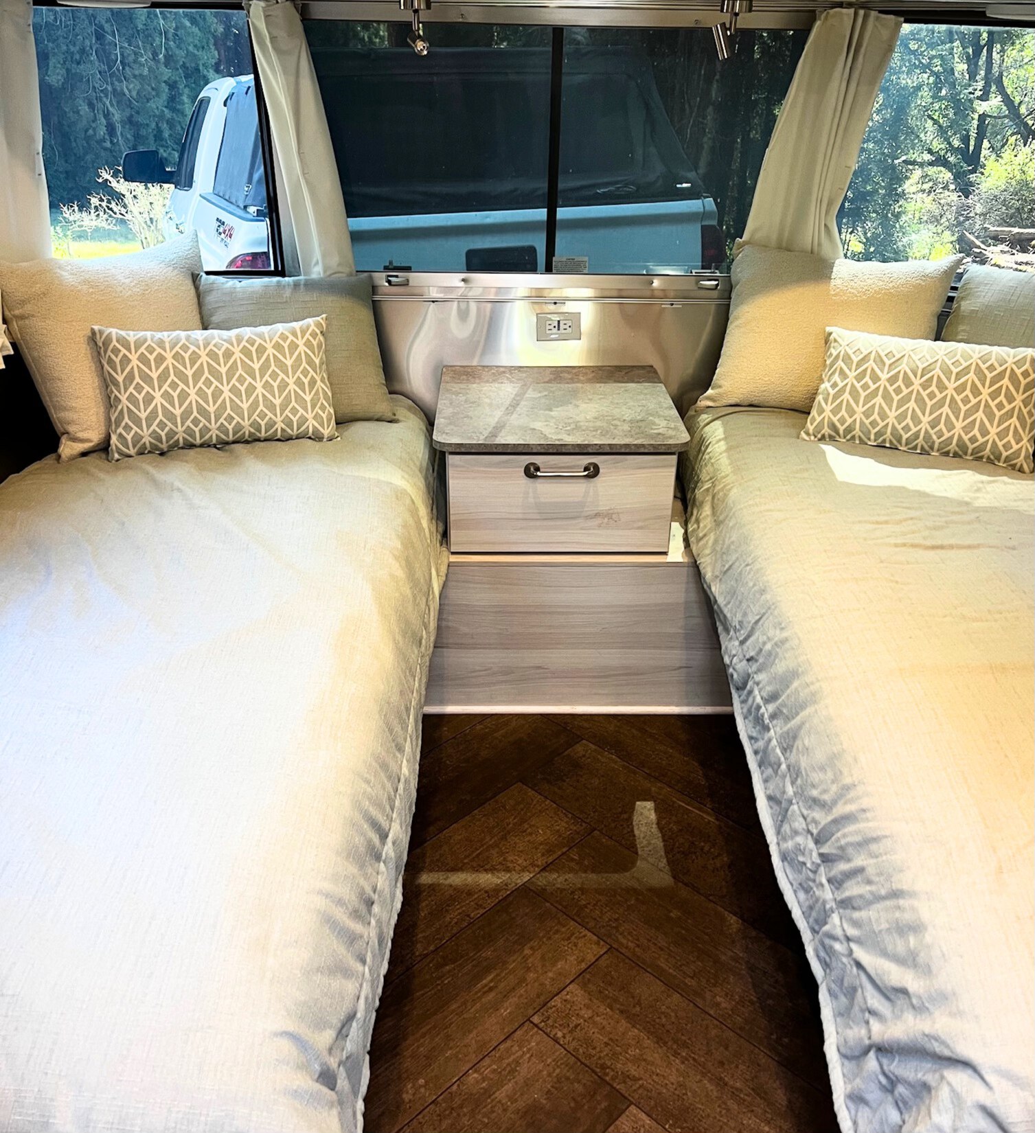 Inside this RV rental, two twin beds with light pillows face each other, separated by a nightstand. Windows show a truck & trees outside.. Airstream International 2025