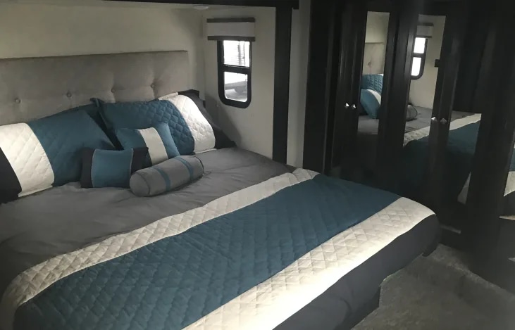 Cozy RV rental bedroom features a comfy grey bed with teal accents, a padded headboard, and mirrored closets.. Heartland RVs Road Warrior Toy Hauler 2017