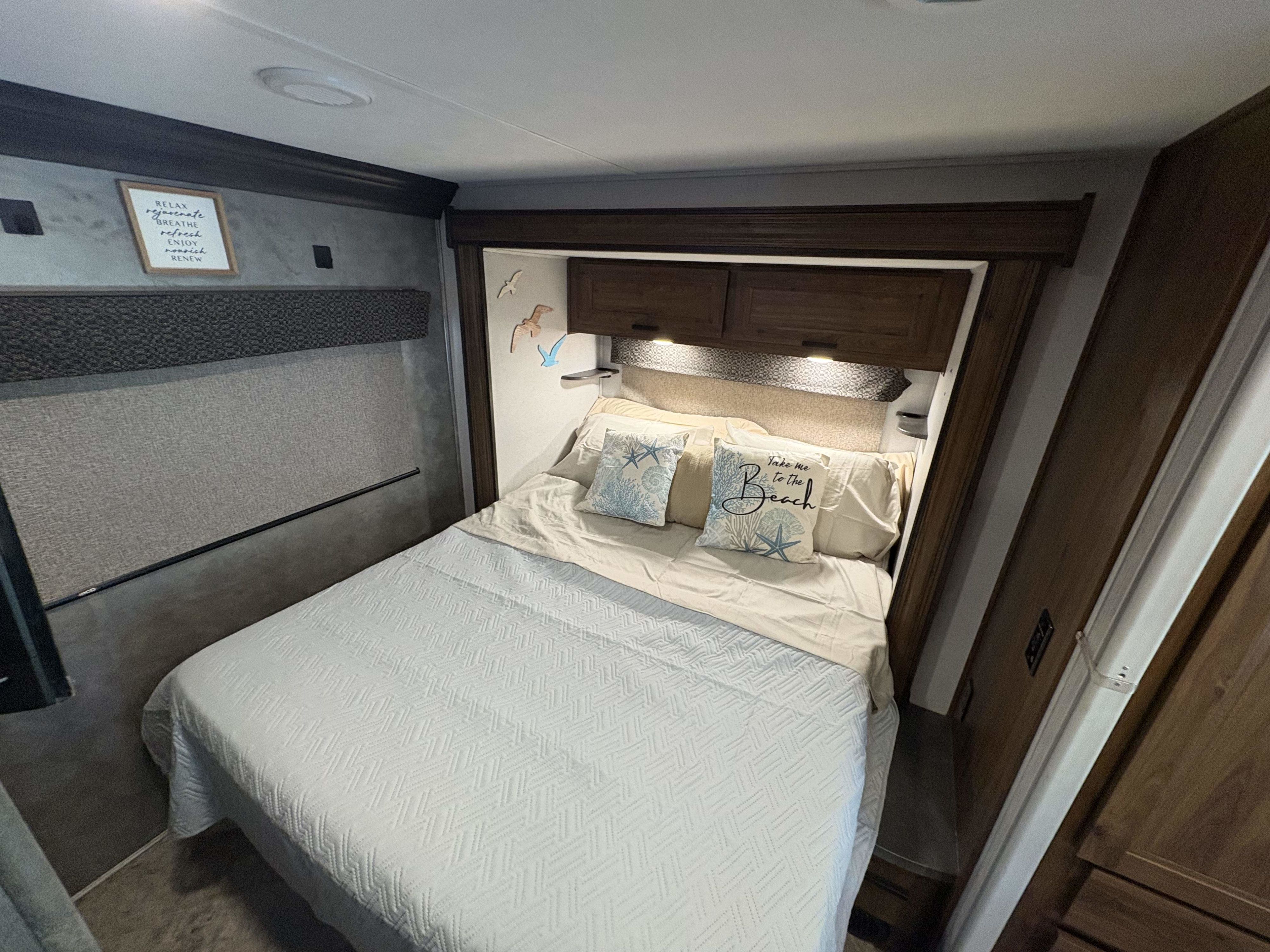 Relaxing RV rental bedroom featuring a comfy bed with coastal pillows & overhead cabinets. Enjoy a restful journey.. Forest River Sunseeker LE 2021
