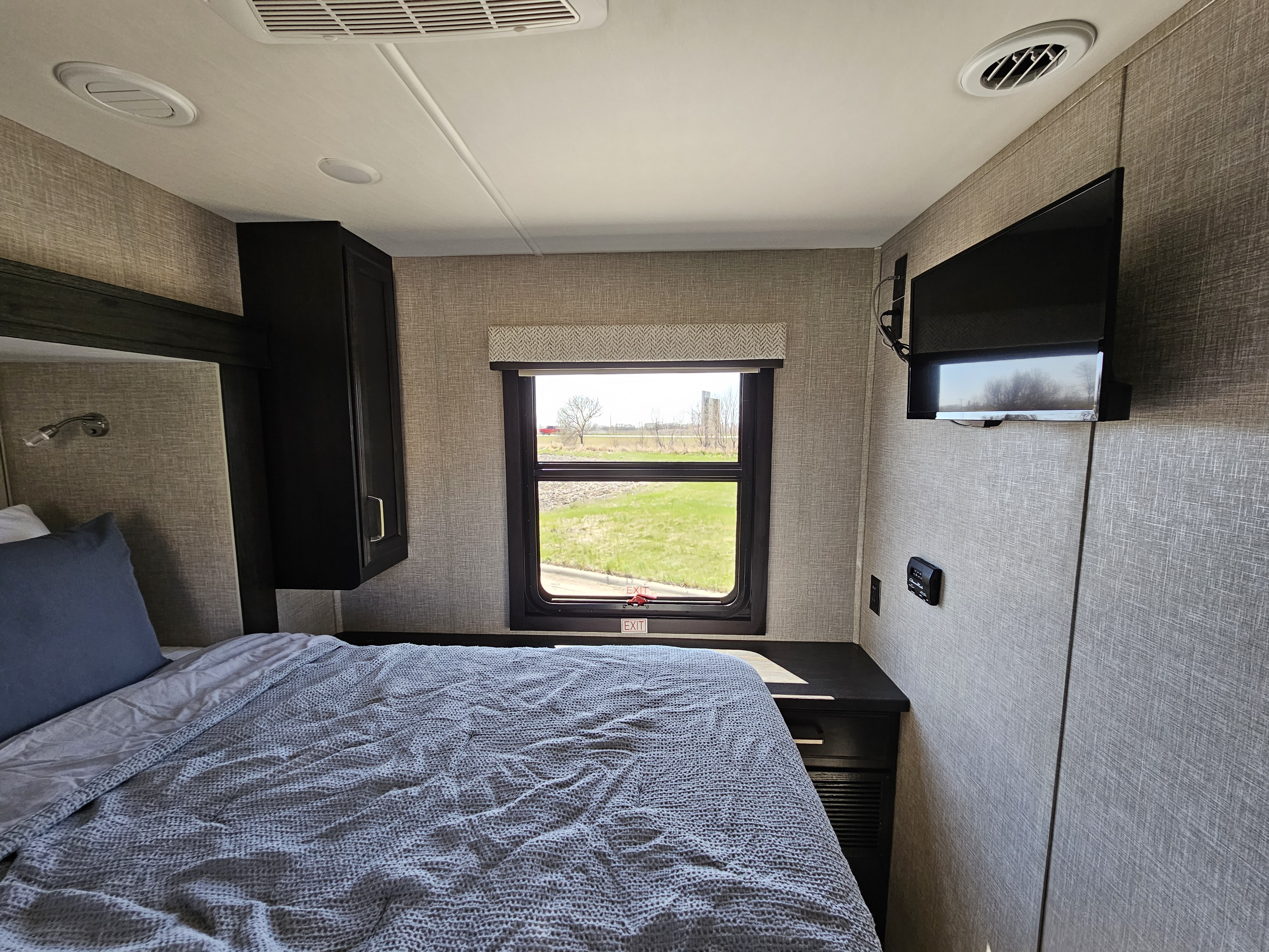 Cozy RV rental bedroom with a comfy bed, window view of green fields, wall-mounted TV, and storage.. Jayco Greyhawk 2023