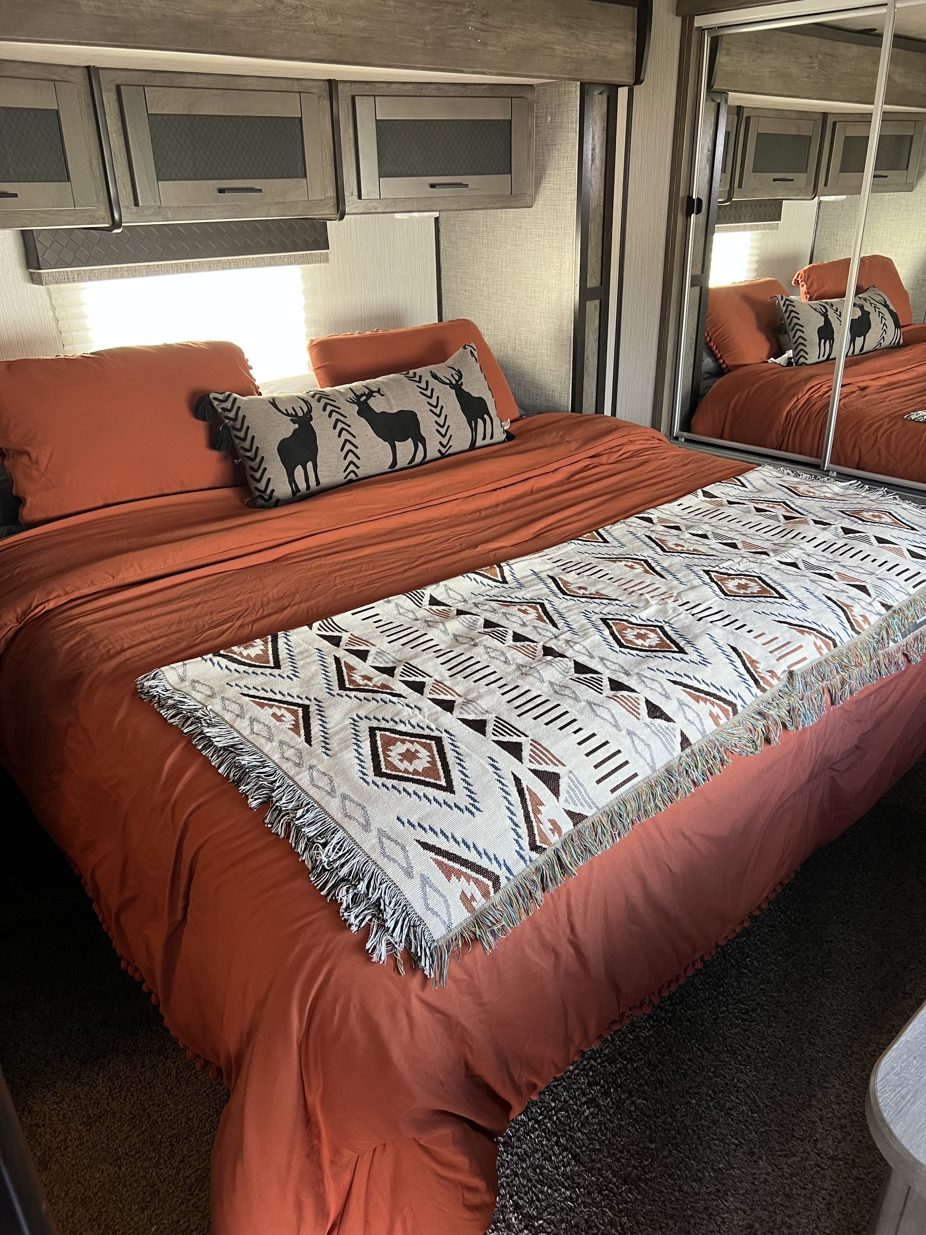 Inviting RV rental bedroom with warm rust-colored bedding, deer pillow, patterned throw, and mirrored closet.. Heartland RVs North Trail 2021