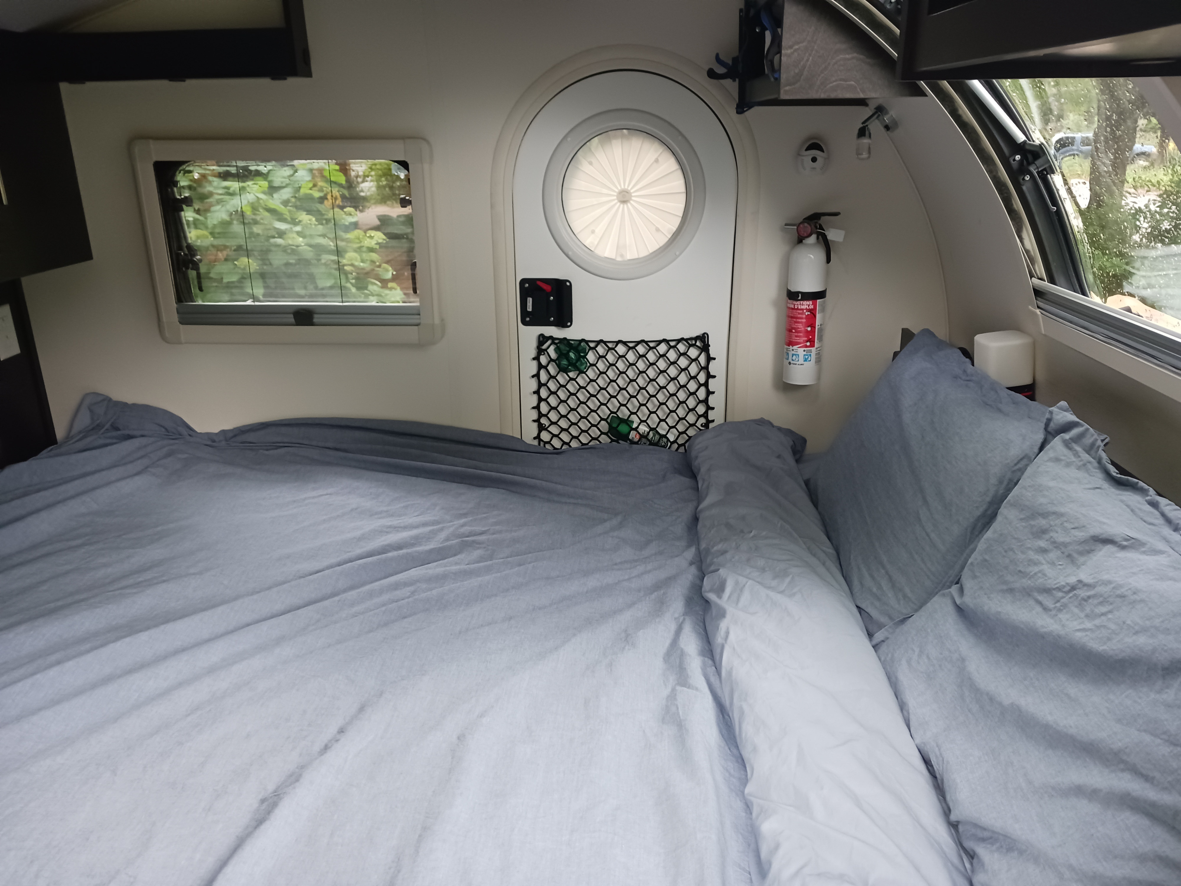 Cozy interior of an RV rental: features a made bed, windows with outdoor views, and a fire extinguisher for safety.. nuCamp T@G 2023