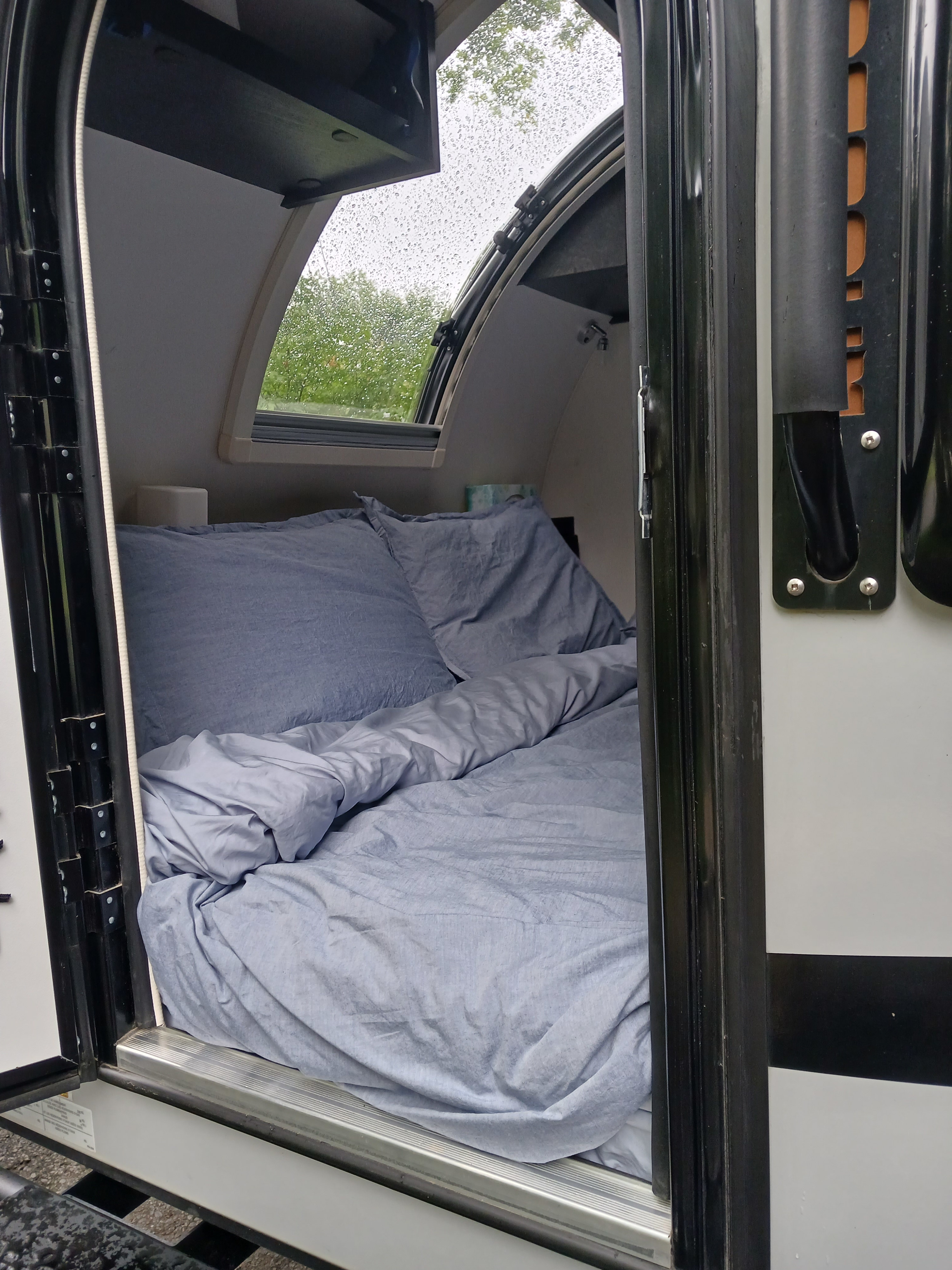 View into a cozy RV rental bedroom. Blue bedding awaits, with raindrops on the window framing green trees outside. Ready for travel!. nuCamp T@G 2023