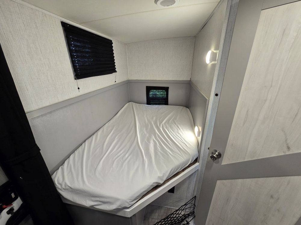 Cozy RV rental sleeping nook: white-sheeted bed, small window with black blind, and bright wall lights on textured walls.. Heartland North Trail 29BHP 2022