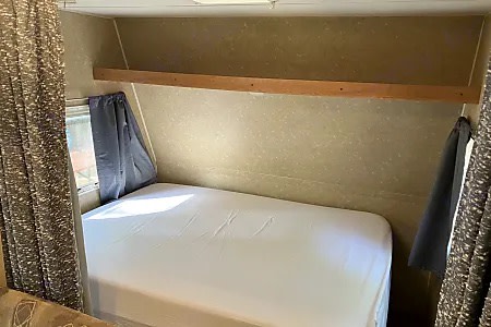 This RV rental features a cozy sleeping area with a white sheet on the bed, a window with curtains, and an overhead shelf.. Gulf Stream Ameri-Lite 2009