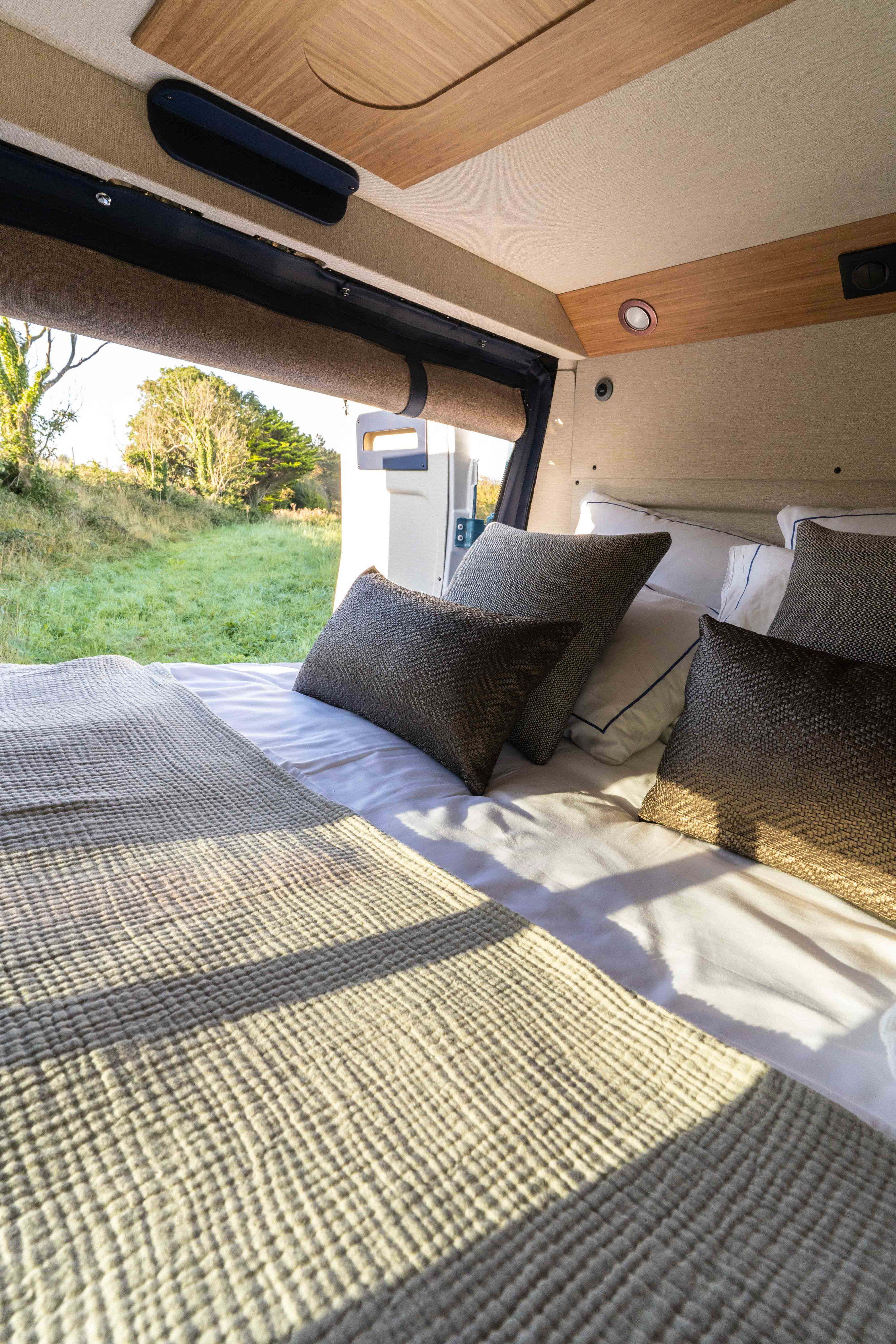 Cozy rv rental interior: A comfortable bed with textured pillows and blanket, offering a direct view of a vibrant grassy field.. Rosalie Type H 2024