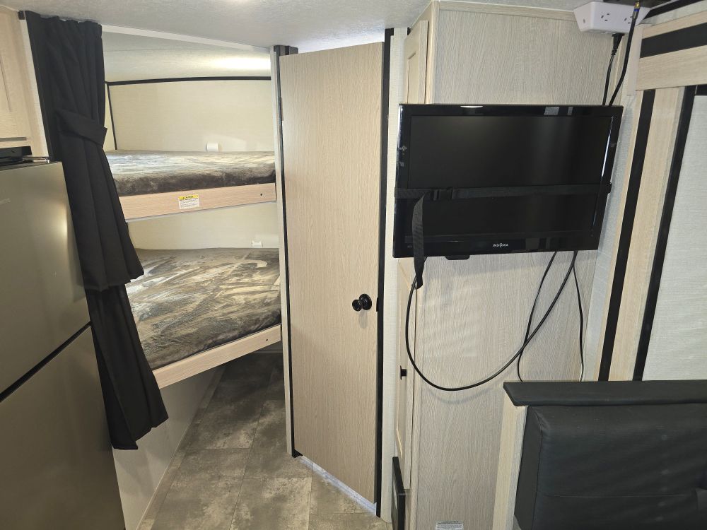RV rental interior with two bunk beds (curtained), a refrigerator, and a mounted Insignia TV, showing compact living.. Coachmen Apex Nano 208BHS 2023