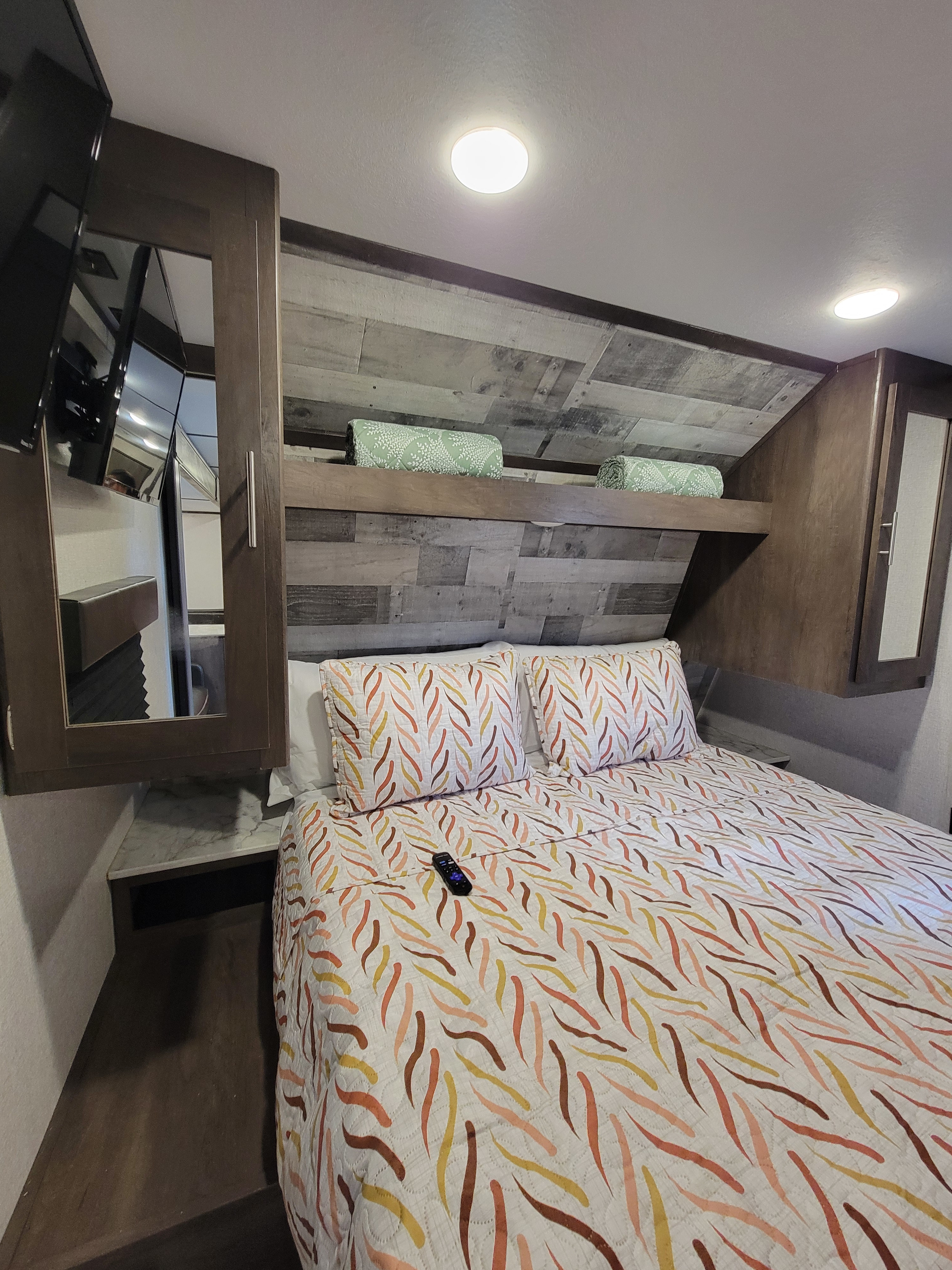 This RV rental bedroom includes a bed with colorful bedding, a wall-mounted TV, and built-in mirrored storage.. Dutchmen Coleman Lantern LT 2021