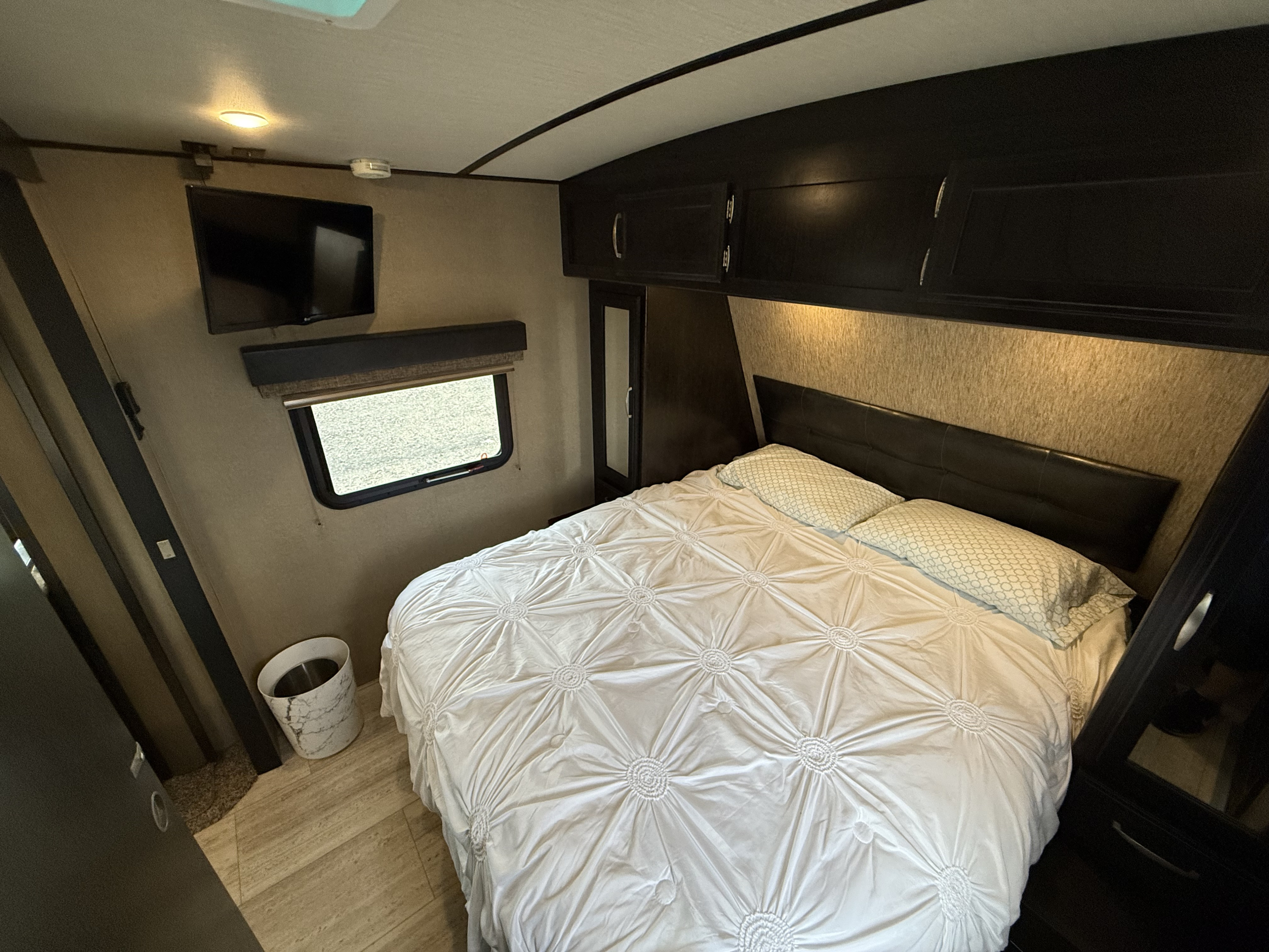 View of a compact RV rental bedroom featuring a large bed, overhead storage cabinets, a wall-mounted TV, and a window.. Grand Design Imagine 2018