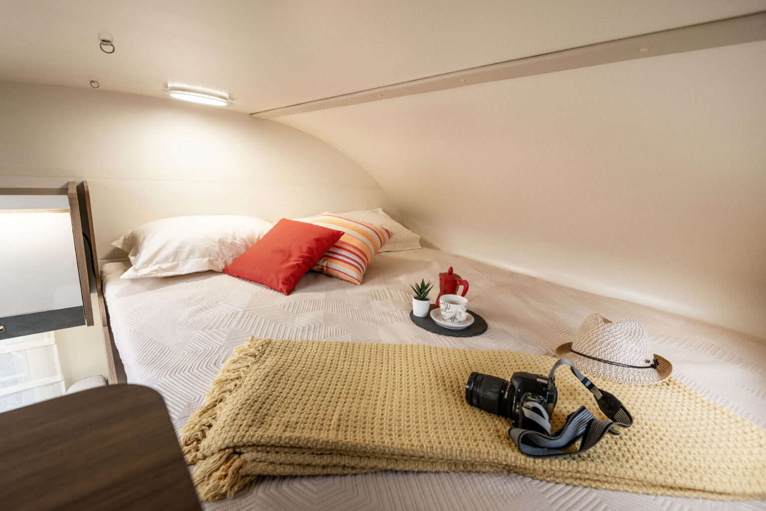 Comfortable bed in an rv rental's sleeping area, adorned with pillows, a blanket, camera, hat, and a coffee setup.. Elnagh baron 20 2025
