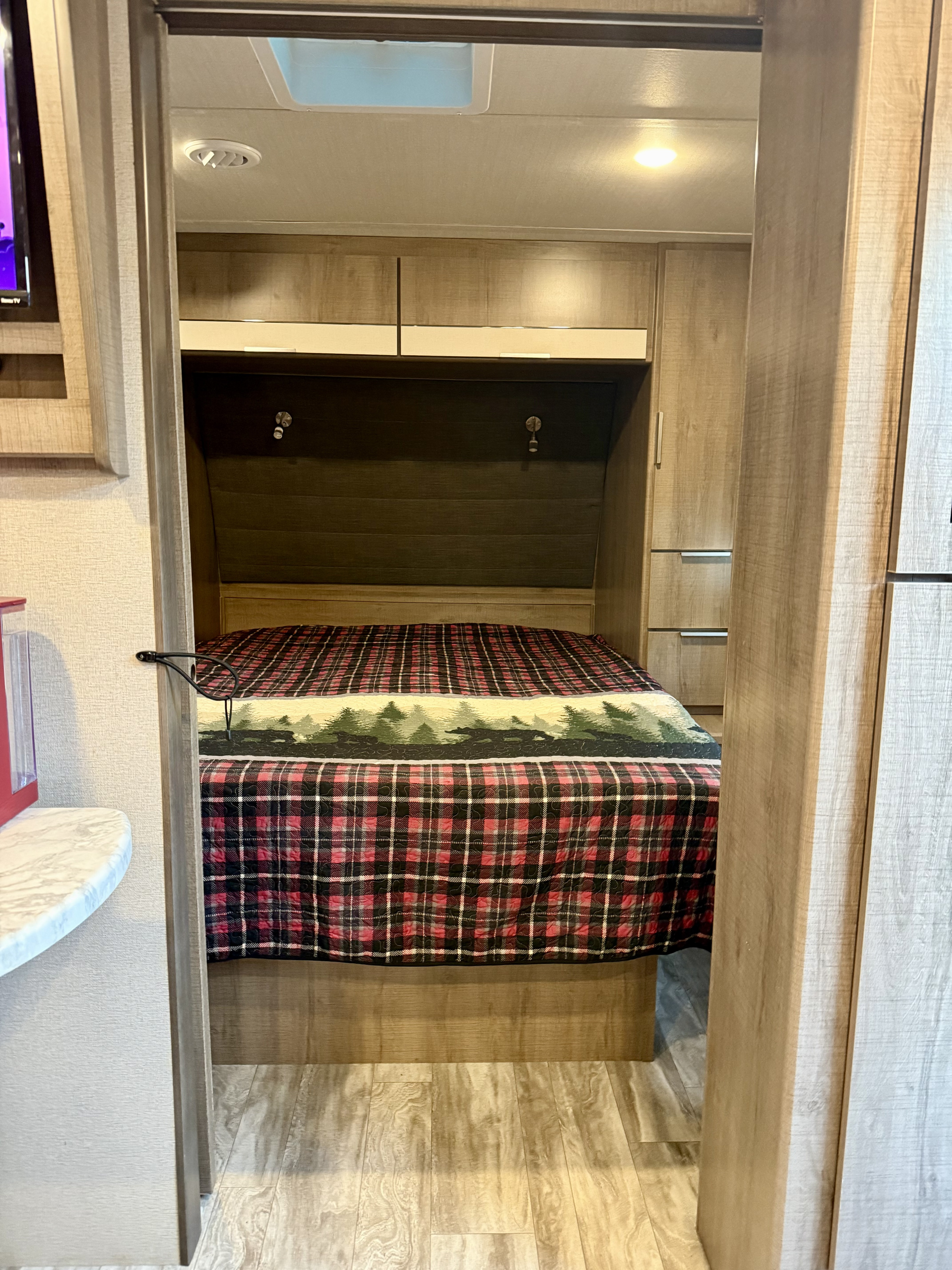 Inside a cozy RV rental bedroom: light wood finishes, plaid comforter with a forest design, and practical storage.. Grand Design Imagine 2021