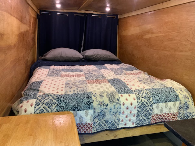 Inside an RV rental, a cozy bed with a blue patchwork quilt, gray pillows, and light wood interior walls provides comfortable accommodations.. Ford transit 2016