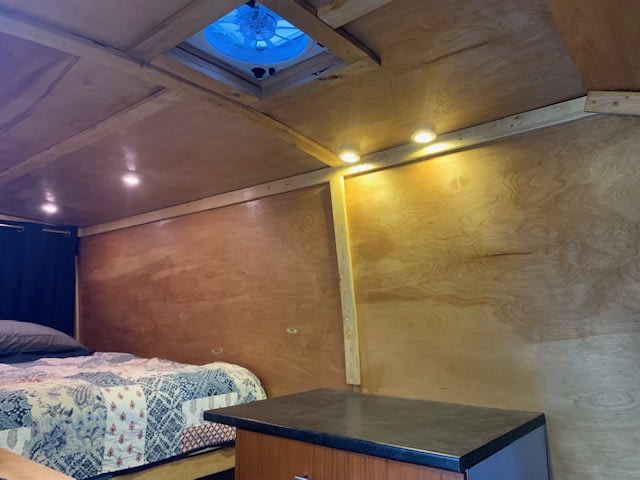 Wood-paneled RV rental interior features a cozy bed, blue ceiling fan, and warm overhead/wall lighting for comfort.. Ford transit 2016