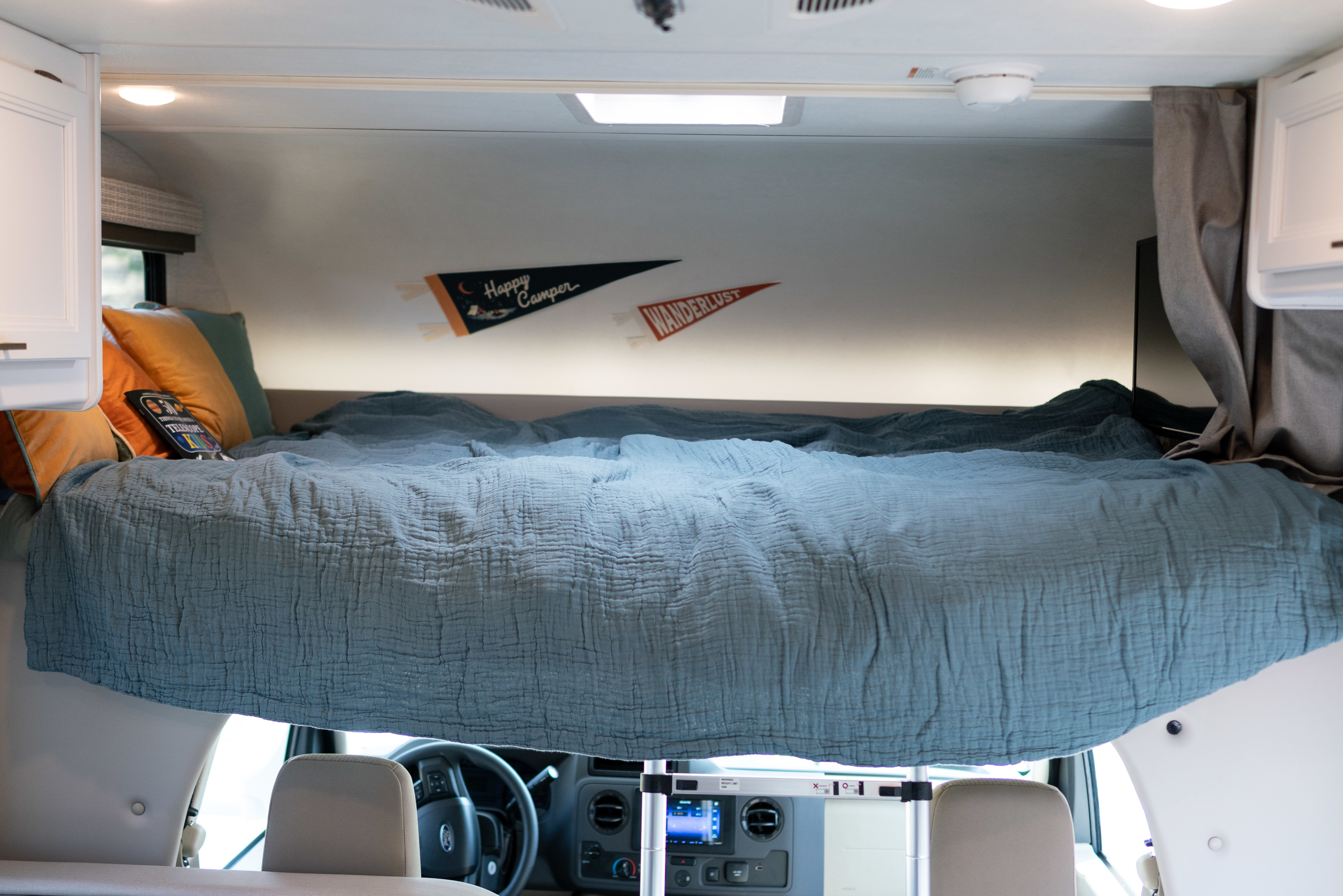 A cozy overhead bed in an RV rental, featuring blue bedding, decorative pillows, and a "Happy Camper" pennant.. Thor Motor Coach Eddie Bauer 21EBF 2025