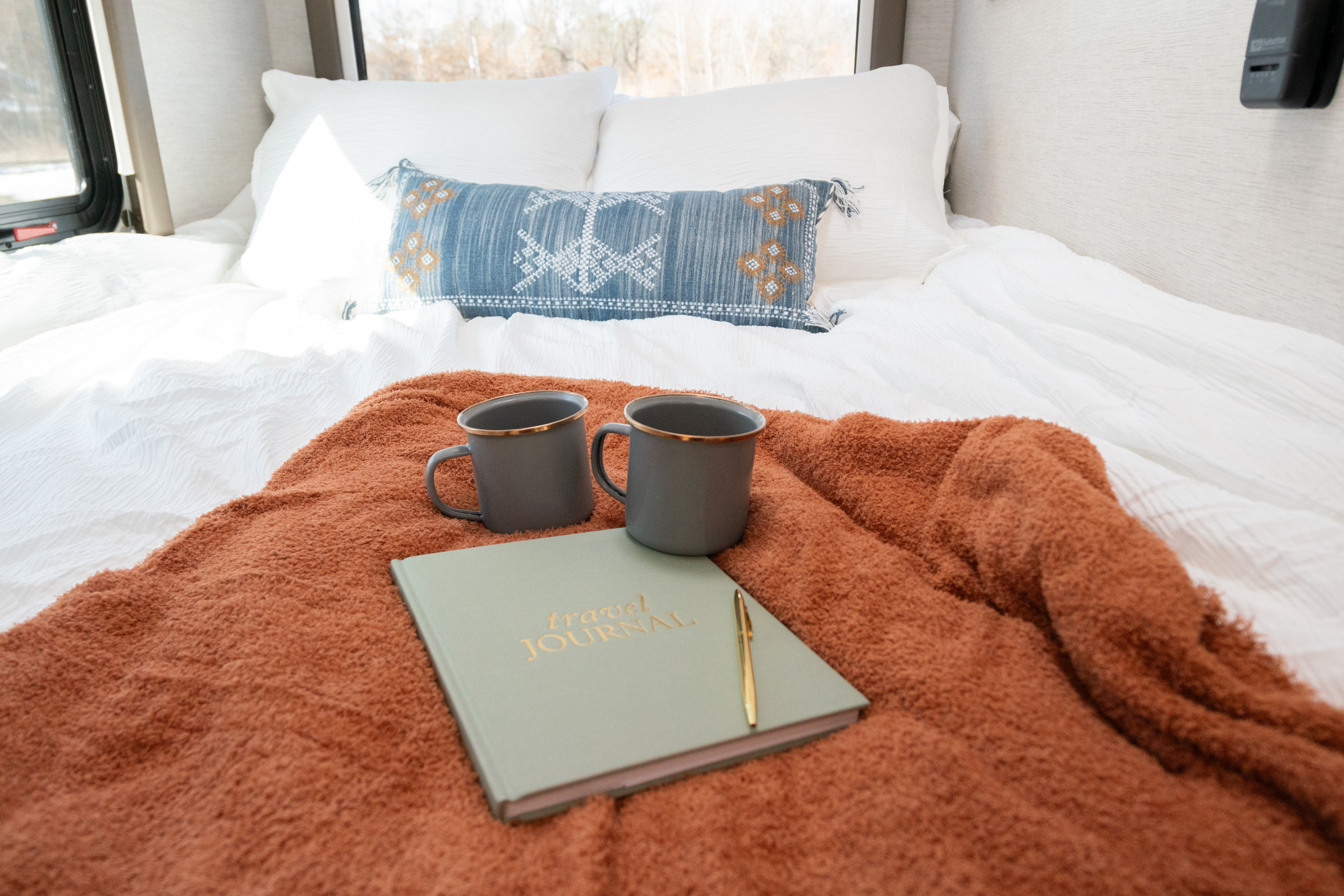 RV rental bed with white bedding, rust plush blanket, grey mugs, and a 'Travel Journal' for cozy adventures.. Thor Motor Coach Eddie Bauer 21EBF 2025