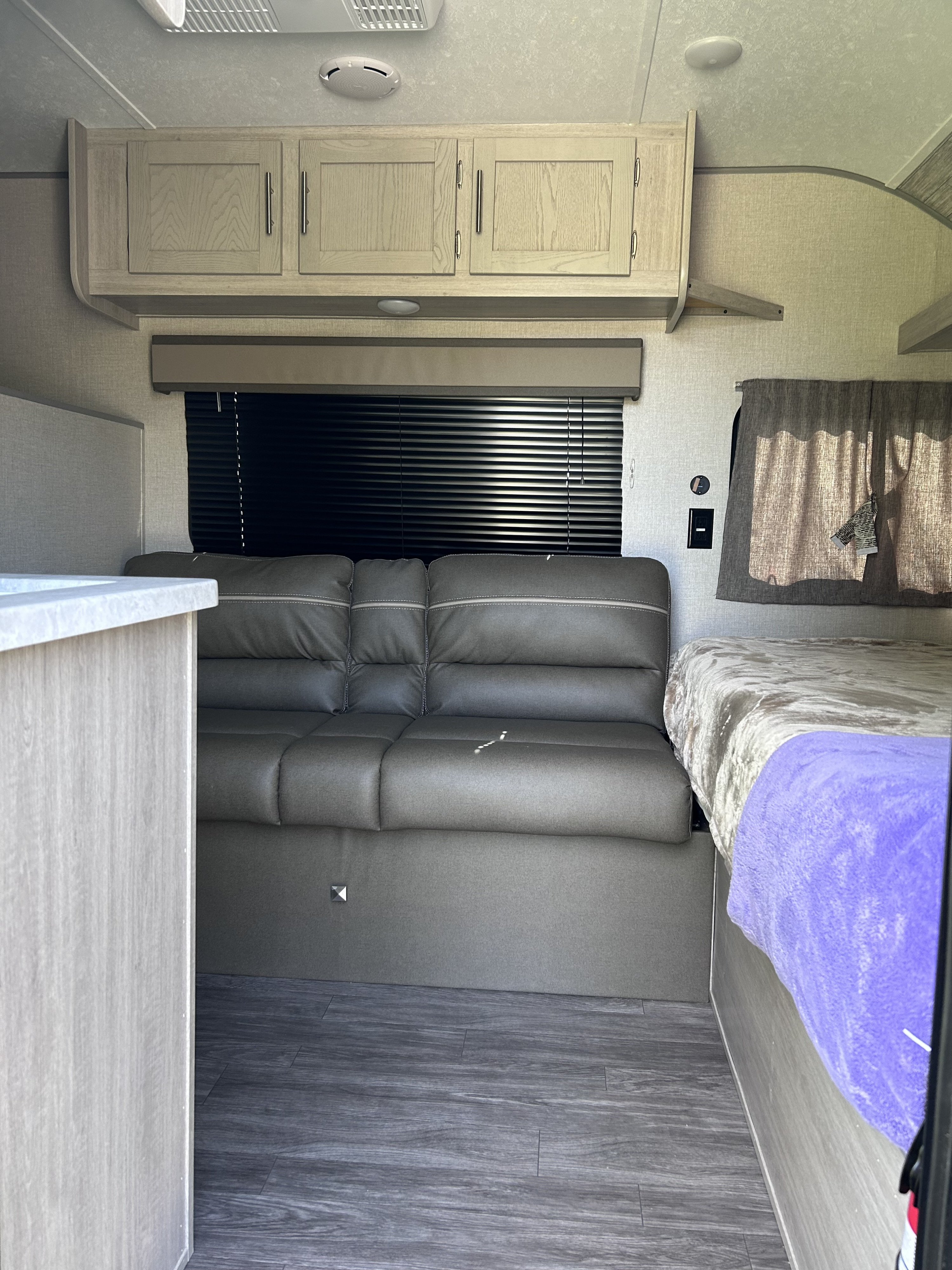 Modern RV rental interior with grey sofa, bed, overhead cabinets, and dark wood-look flooring, showing the living space.. Forest River Aurora Aurora 2023