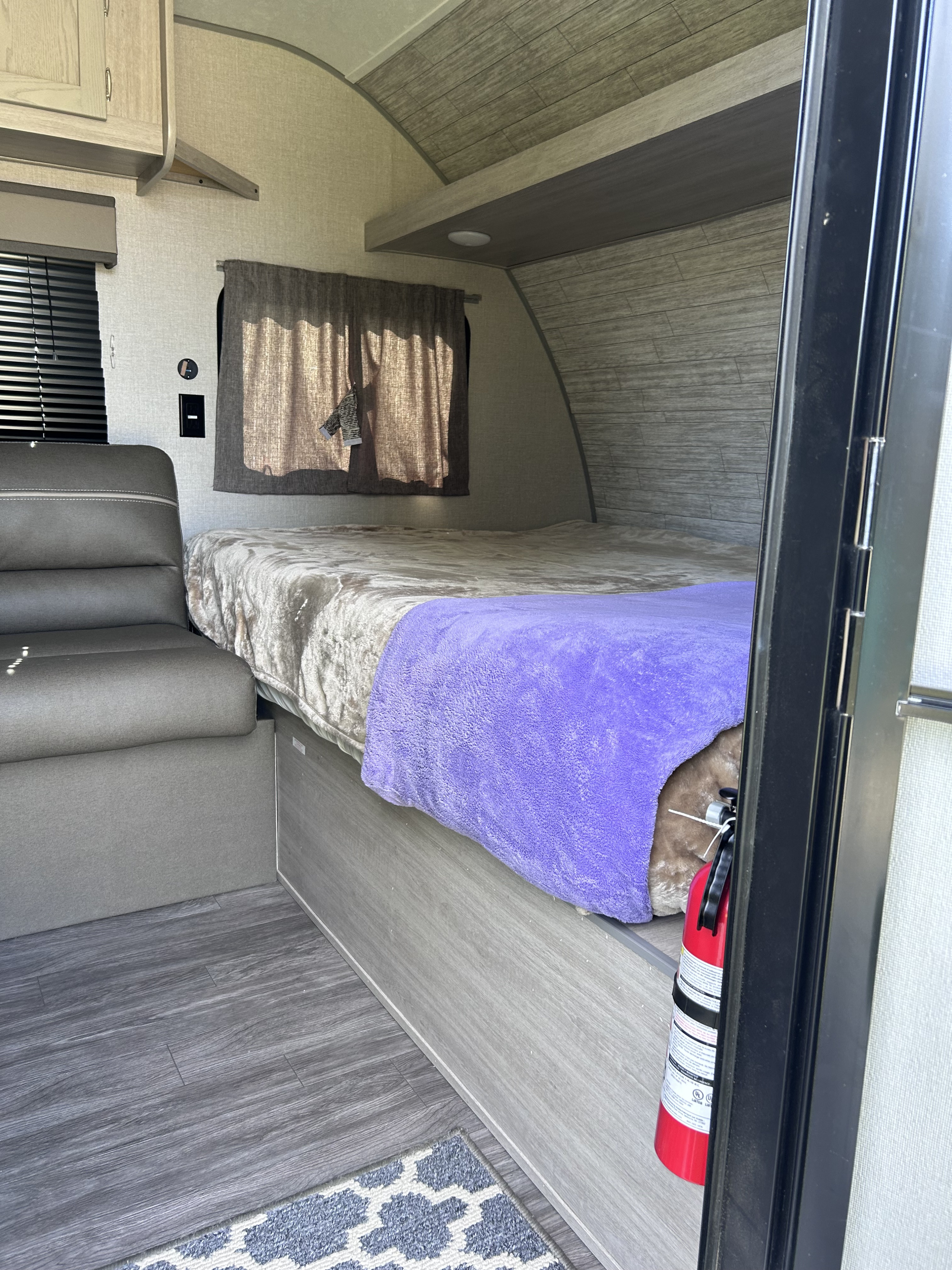 Inside a compact RV rental: a bed with purple throw, gray seating, and wood-look flooring. Fire extinguisher visible.. Forest River Aurora Aurora 2023