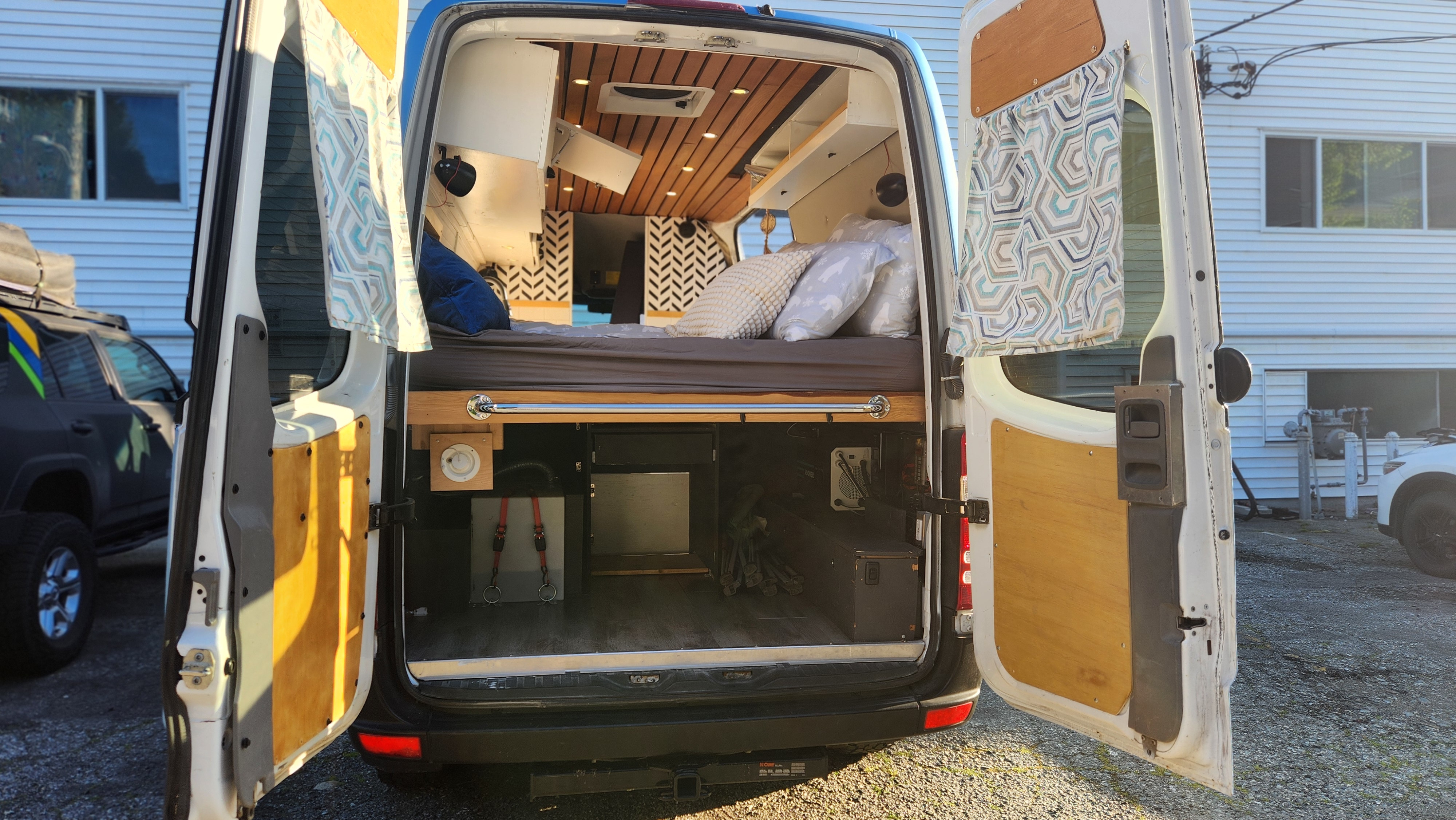 Open doors reveal an RV rental campervan's rear: cozy bed, wood ceiling, and smart under-bed storage for adventures.. Mercedes-Benz Sprinter 2011